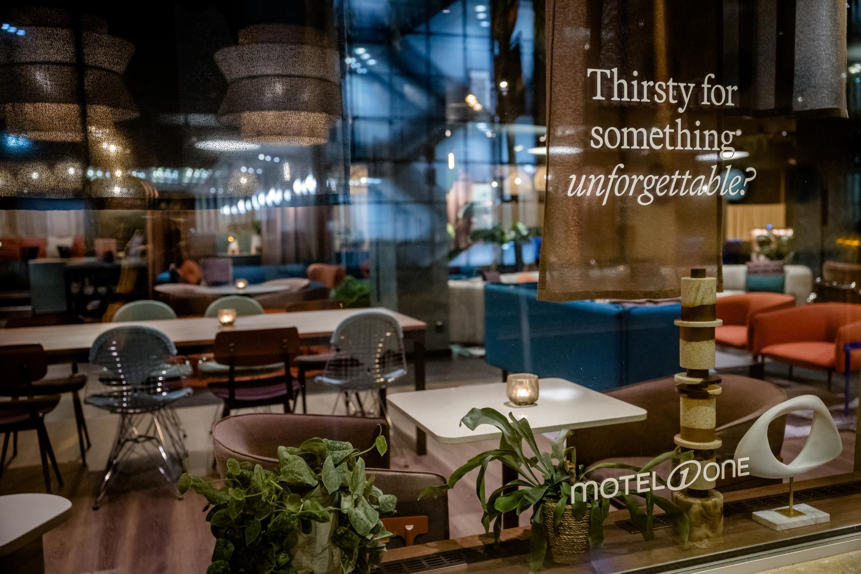 Moderne Hotelbar mit stilvoller Beleuchtung, gemütlichen Sitzbereichen und Pflanzen, einladend für entspannte Momente und besondere Erlebnisse. Schriftzug „Thirsty for something unforgettable?“ sichtbar.