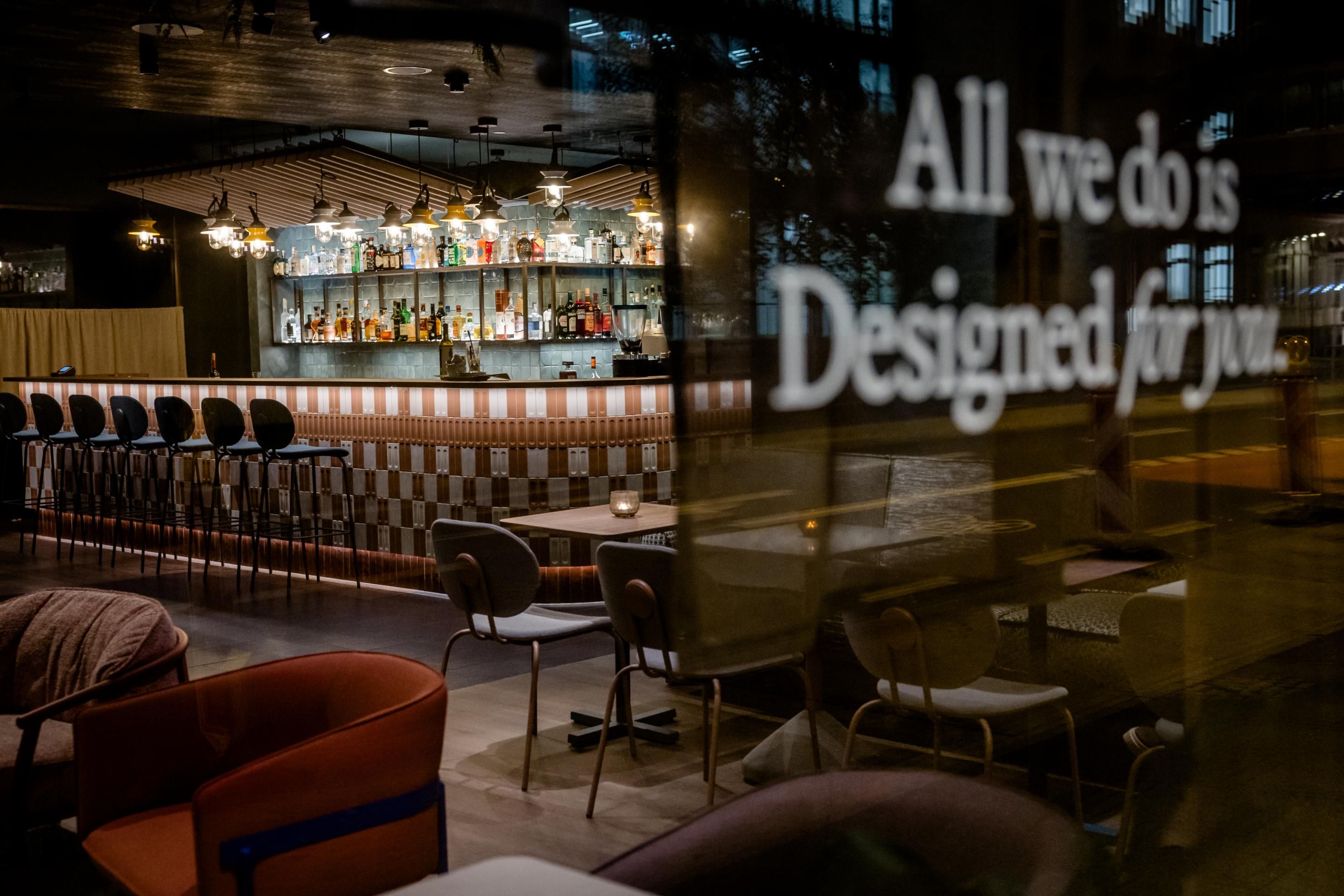 Moderne Hotelbar in warmem Licht, mit gemütlichen Sitzgelegenheiten und einer gut bestückten Getränketheke, die zu entspannten Abenden einlädt. Sichtbar ist der Schriftzug „All we do is Designed for you“ als Aufmerksamkeit auf Gastfreundschaft.