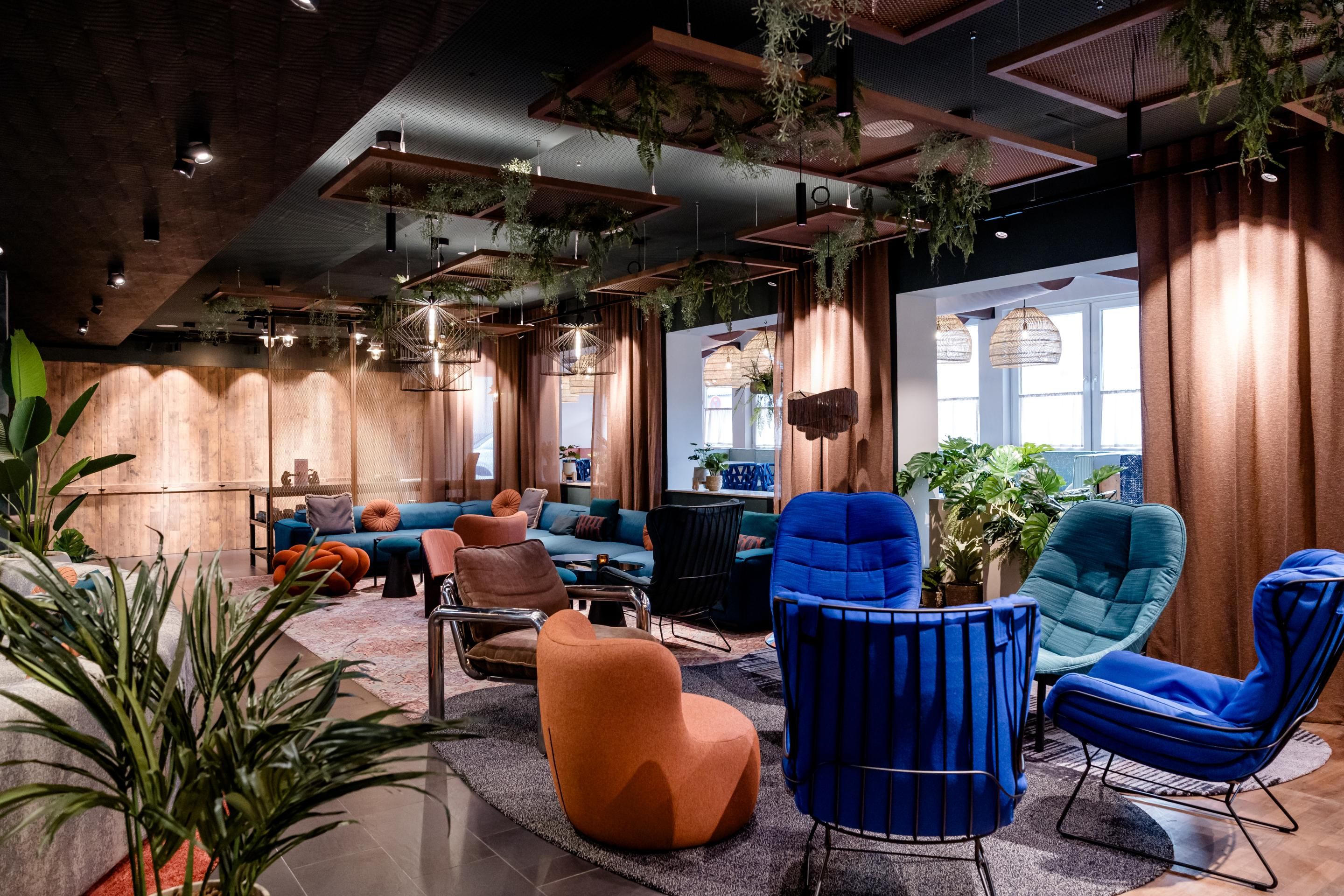Gemütlicher, moderner Lounge-Bereich mit bunten Sesseln, Sofas und Pflanzen, warmem Licht und großen Fenstern, ideal zum Entspannen oder geselligen Beisammensein in einer einladenden Atmosphäre.
