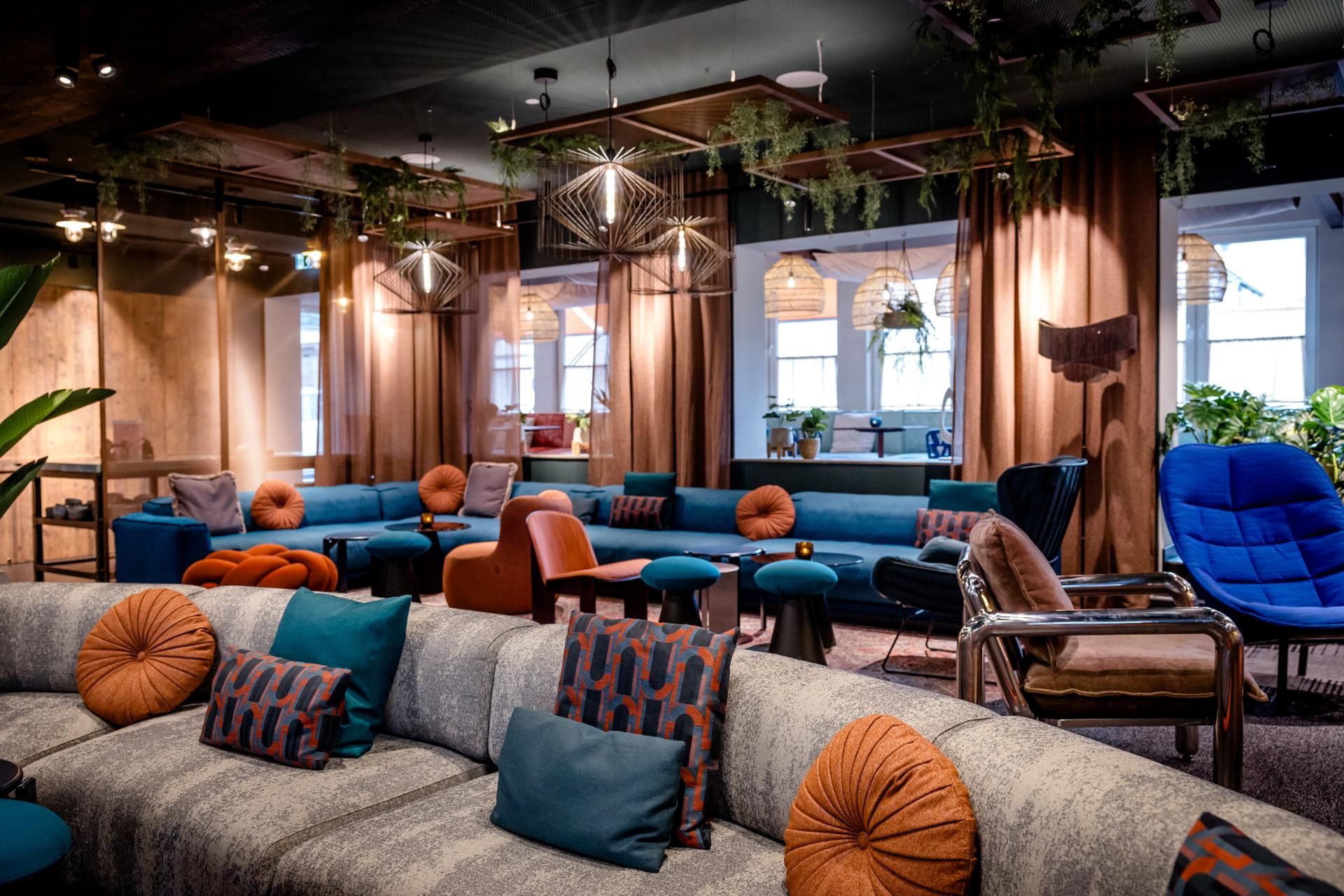 Moderne Hotel-Lounge mit gemütlichen Sofas, farblich abgestimmten Kissen und warmem Licht. Pflanzen und große Fenster schaffen eine einladende, entspannte Atmosphäre zum Verweilen und Austausch.