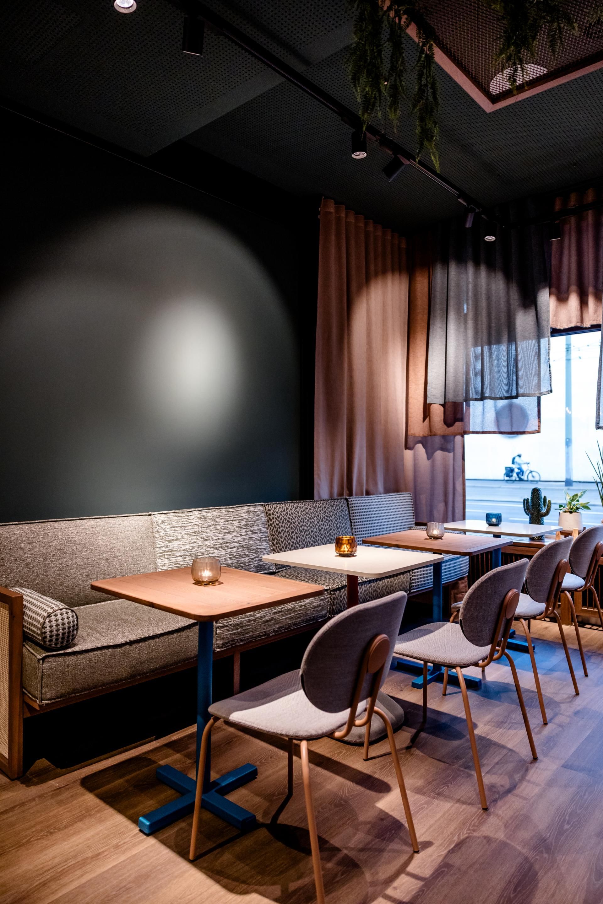 Gemütlicher Innenraum eines Restaurants mit modernen Holztischen, Stühlen und einer gepolsterten Sitzbank, weiche Vorhänge und gedämpftes Licht schaffen eine entspannte Atmosphäre zum Essen und Verweilen.