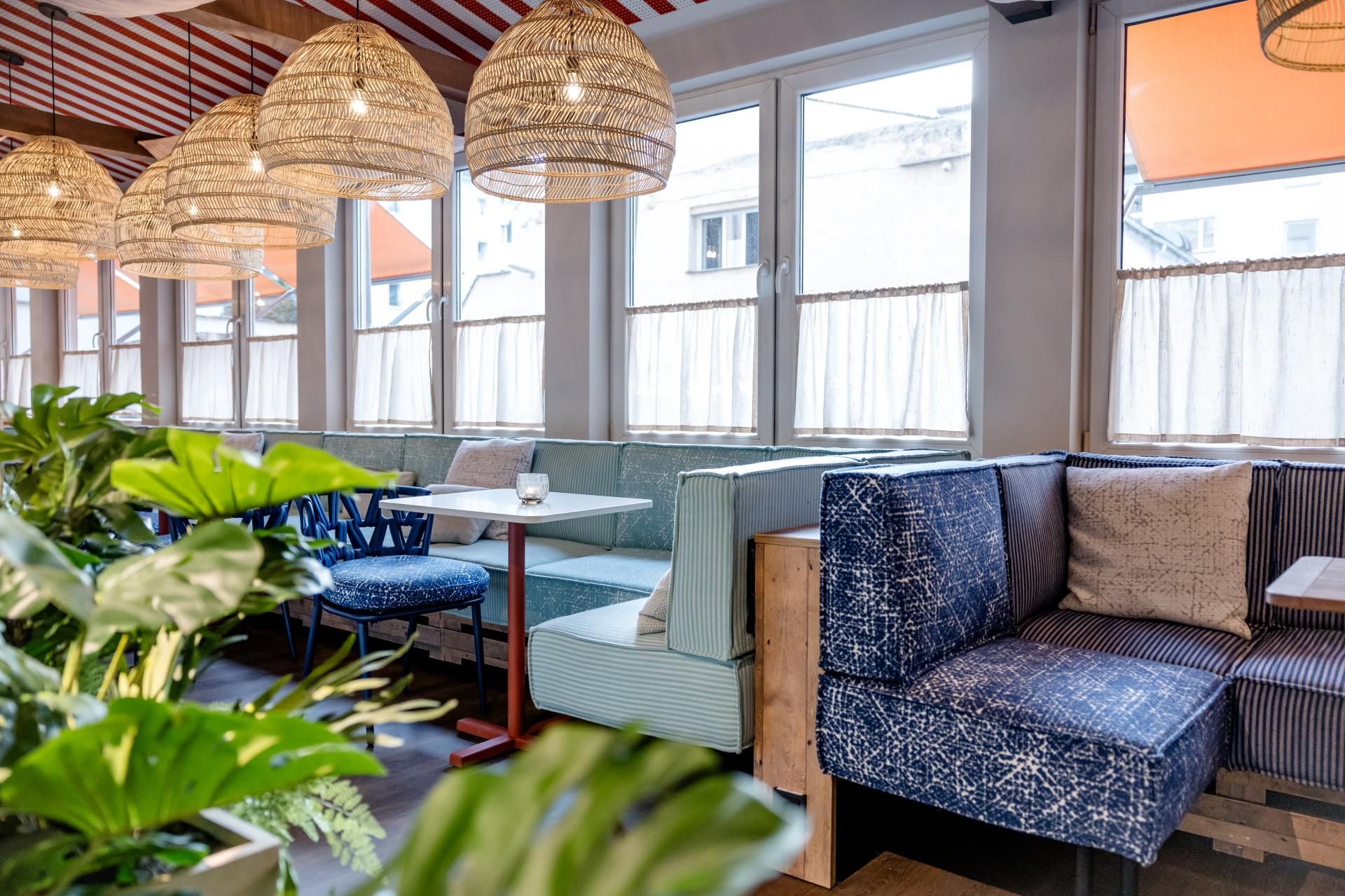 Gemütliche Lounge-Ecke mit gepolsterten Sitzbänken, Dekokissen und großen Fenstern, durch die Tageslicht fällt. Natürliche Pflanzen und warme, geflochtene Hängeleuchten schaffen eine einladende Atmosphäre zum Entspannen oder geselligen Beisammensein.