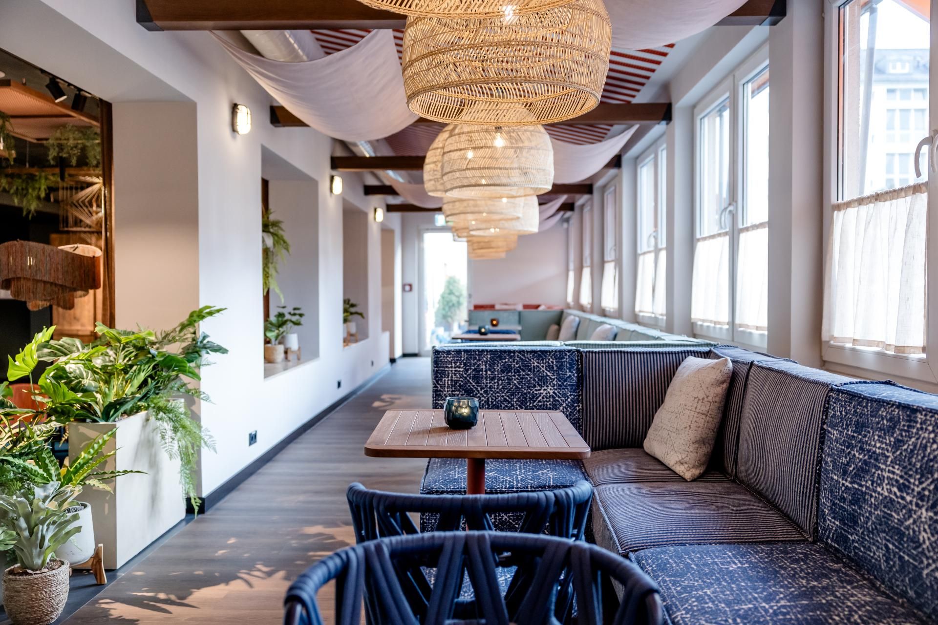 Gemütliche Lounge mit runden Deckenlampen, hellen Fenstern und gepolsterten Sitzbänken. Natürliche Dekoration mit Pflanzen schafft entspannte Atmosphäre zum Verweilen.