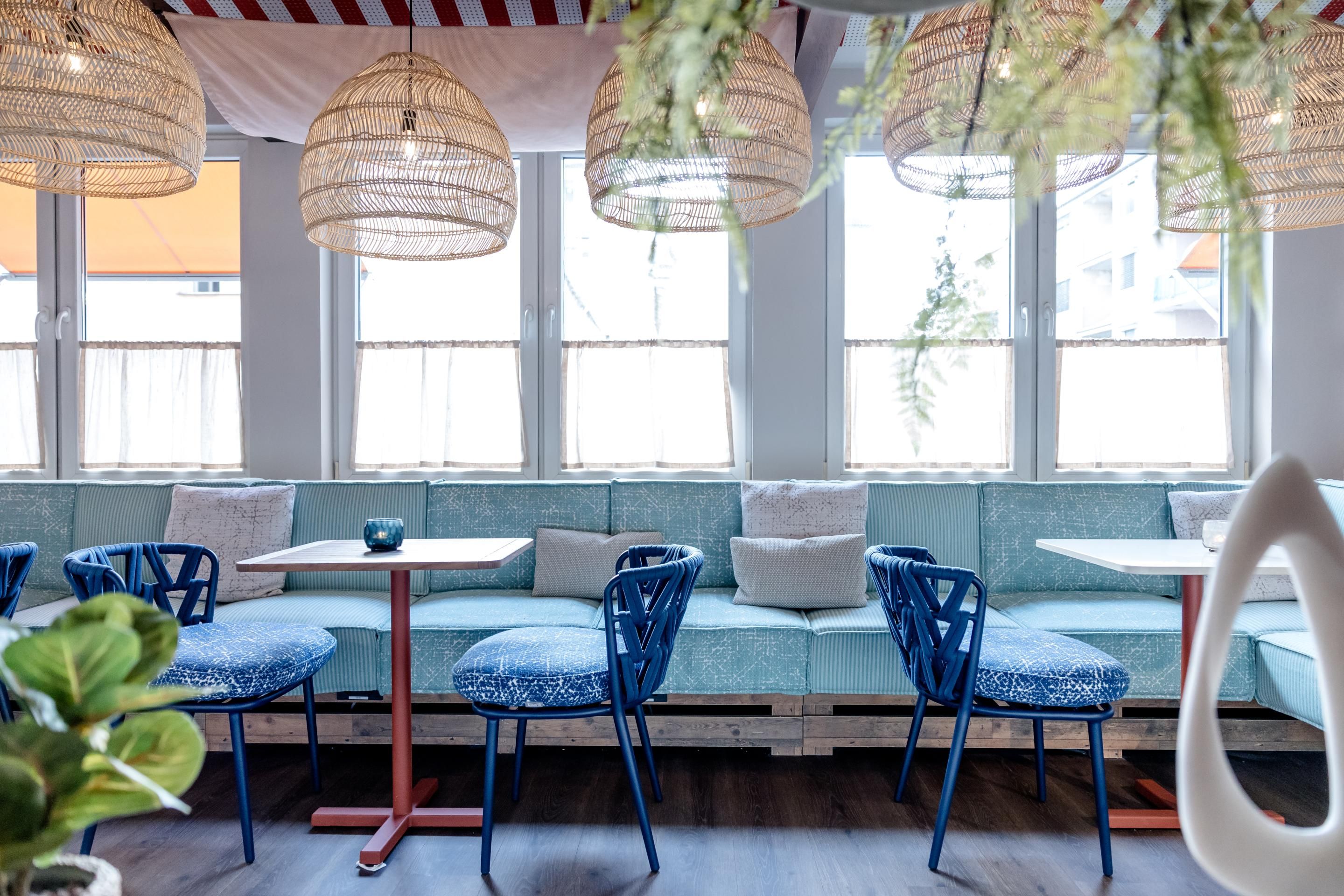 Helles, modernes Café mit großen Fenstern, blauen Stühlen, hellblauen Polstersofas und natürlichen Korbhängelampen, das eine einladende und entspannte Atmosphäre schafft.