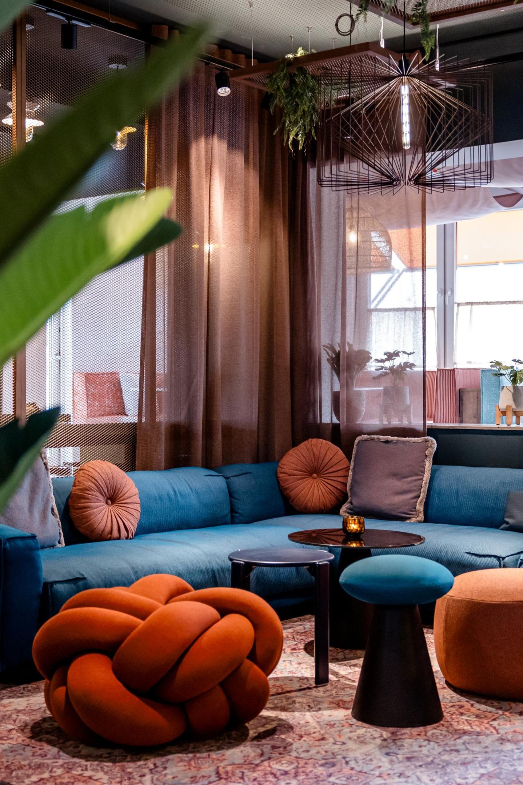 Gemütlicher Loungeraum mit blauem Ecksofa, bunten runden Kissen und stilvollen Sesseln in warmen Erdtönen, umgeben von bodentiefen Vorhängen und Pflanzen, der eine entspannte Atmosphäre für Gäste schafft.