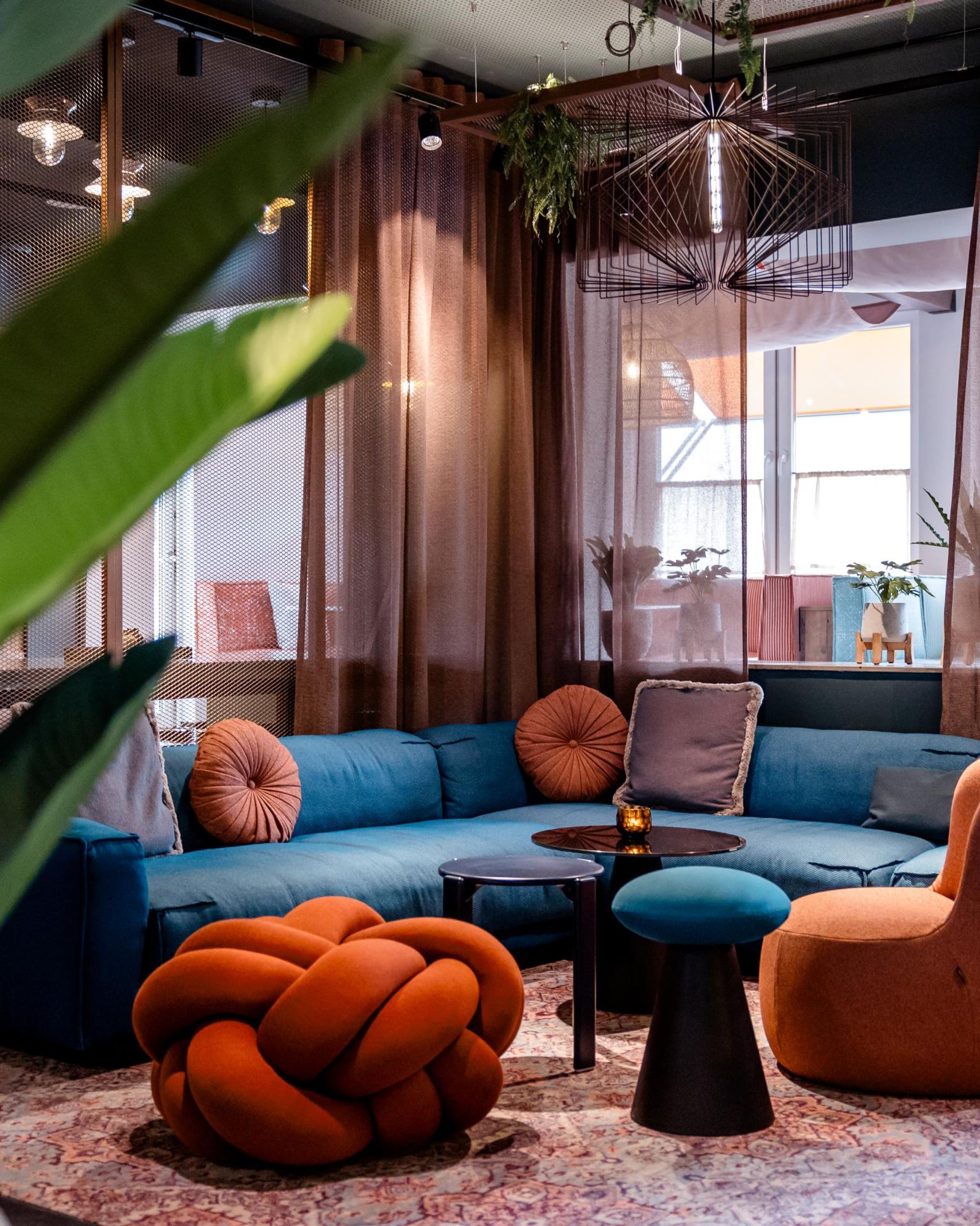 Gemütlicher Loungeraum mit blauem Ecksofa, bunten runden Kissen und stilvollen Sesseln in warmen Erdtönen, umgeben von bodentiefen Vorhängen und Pflanzen, der eine entspannte Atmosphäre für Gäste schafft.