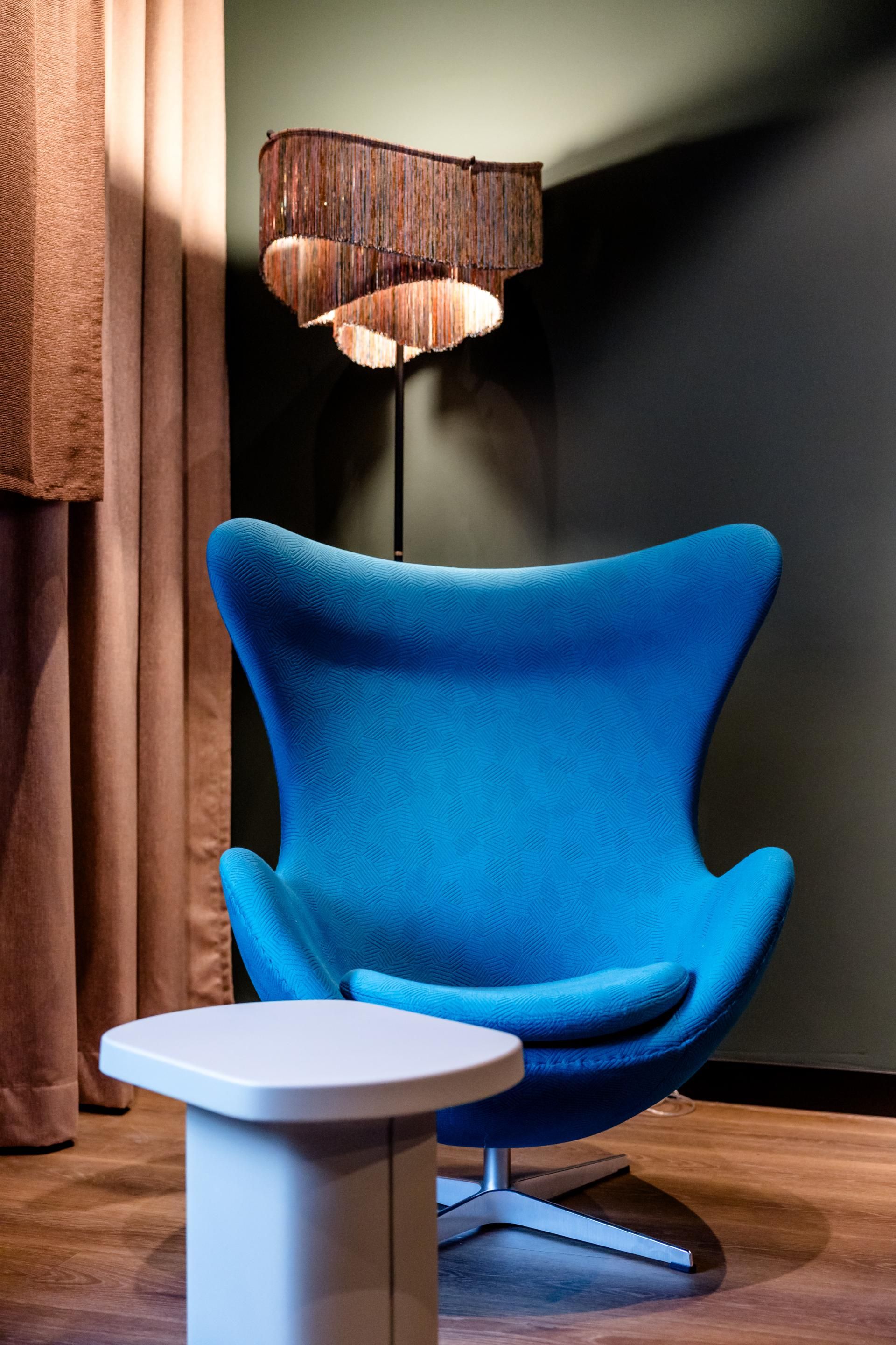 Moderner, bequemer blauer Loungesessel vor dunkler Wand, ergänzt durch einen kleinen weißen Tisch und eine dekorative Stehlampe mit Fransen, schafft eine gemütliche Sitzecke zum Entspannen.