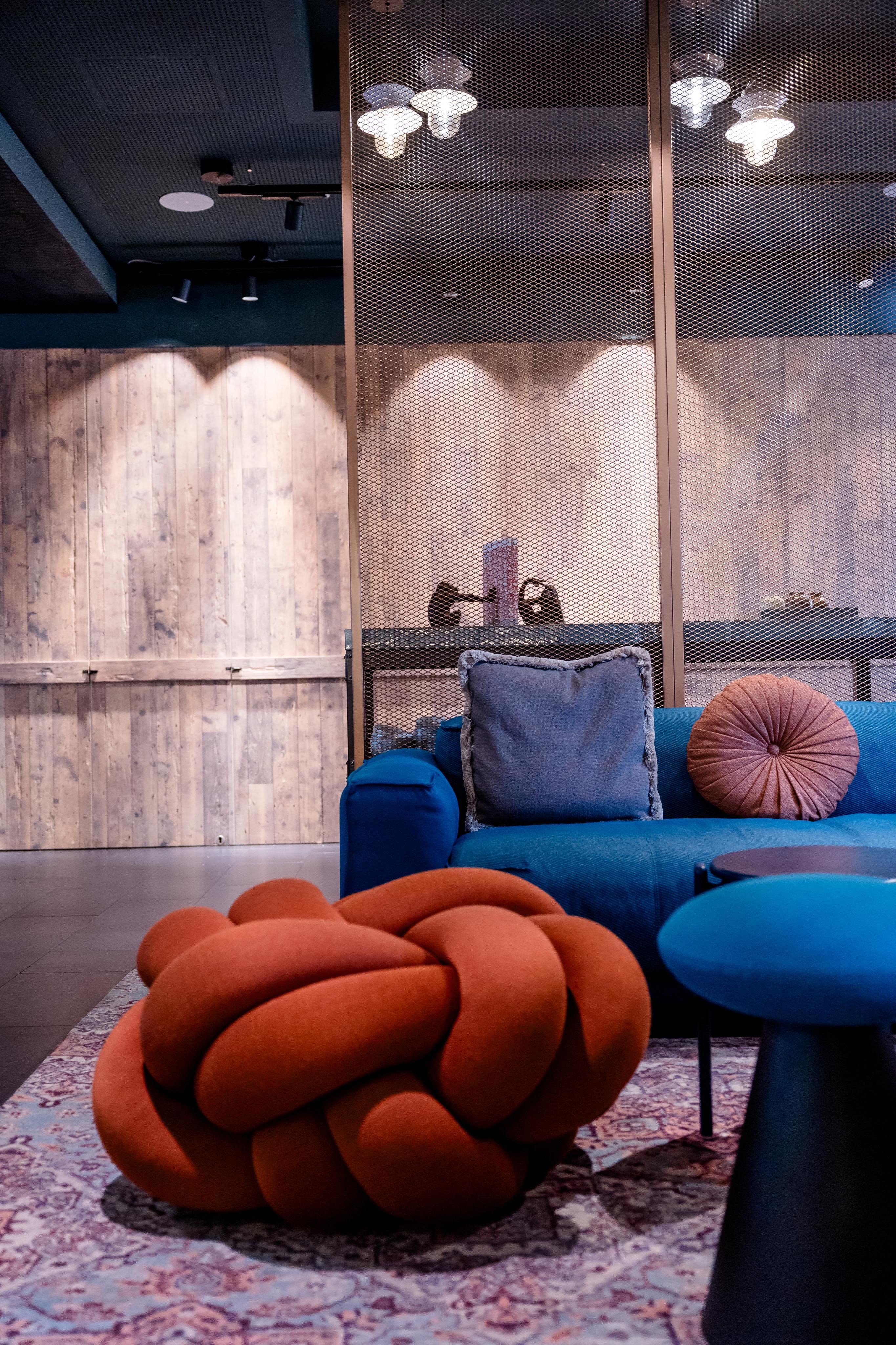 Moderner Lounge-Bereich mit blauer Couch, dekorativen Kissen und einem auffälligen, orangefarbenen Knotenhocker auf Teppichboden vor Holzpaneelwand, geschaffen für entspanntes Verweilen in stilvollem Ambiente.