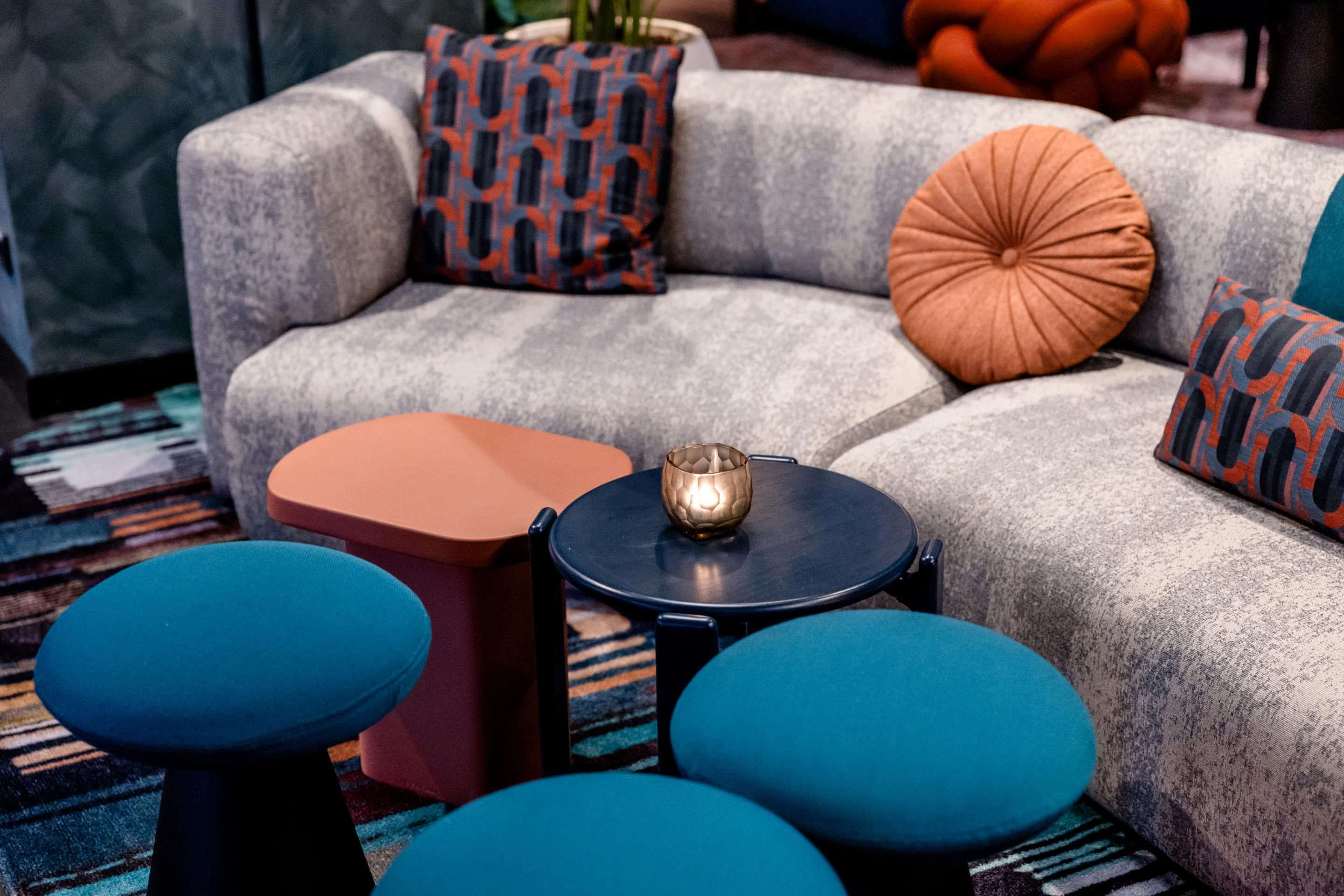 Gemütliche Sitzecke mit grauem Sofa, bunten Kissen und runden, blauen Hockern, dazu kleine Tische mit dekorativer Kerze – einladendes Ambiente zum Entspannen und Verweilen.