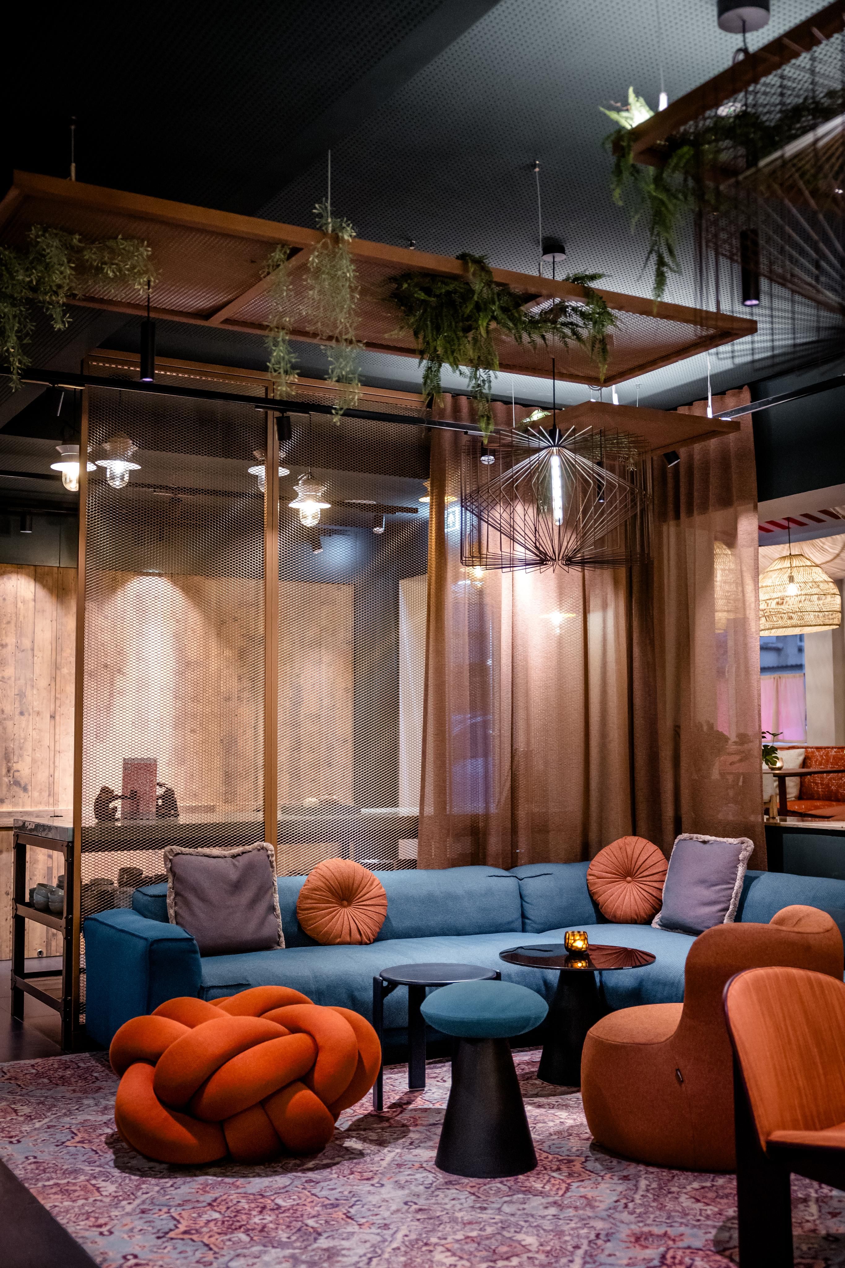 Gemütliche Lounge mit blauem Sofa, orangen und lilafarbenen Kissen, modernem Teppich und überhängenden Pflanzen. Warme Beleuchtung und stilvolle Sitzmöbel schaffen eine entspannte Atmosphäre zum Verweilen.