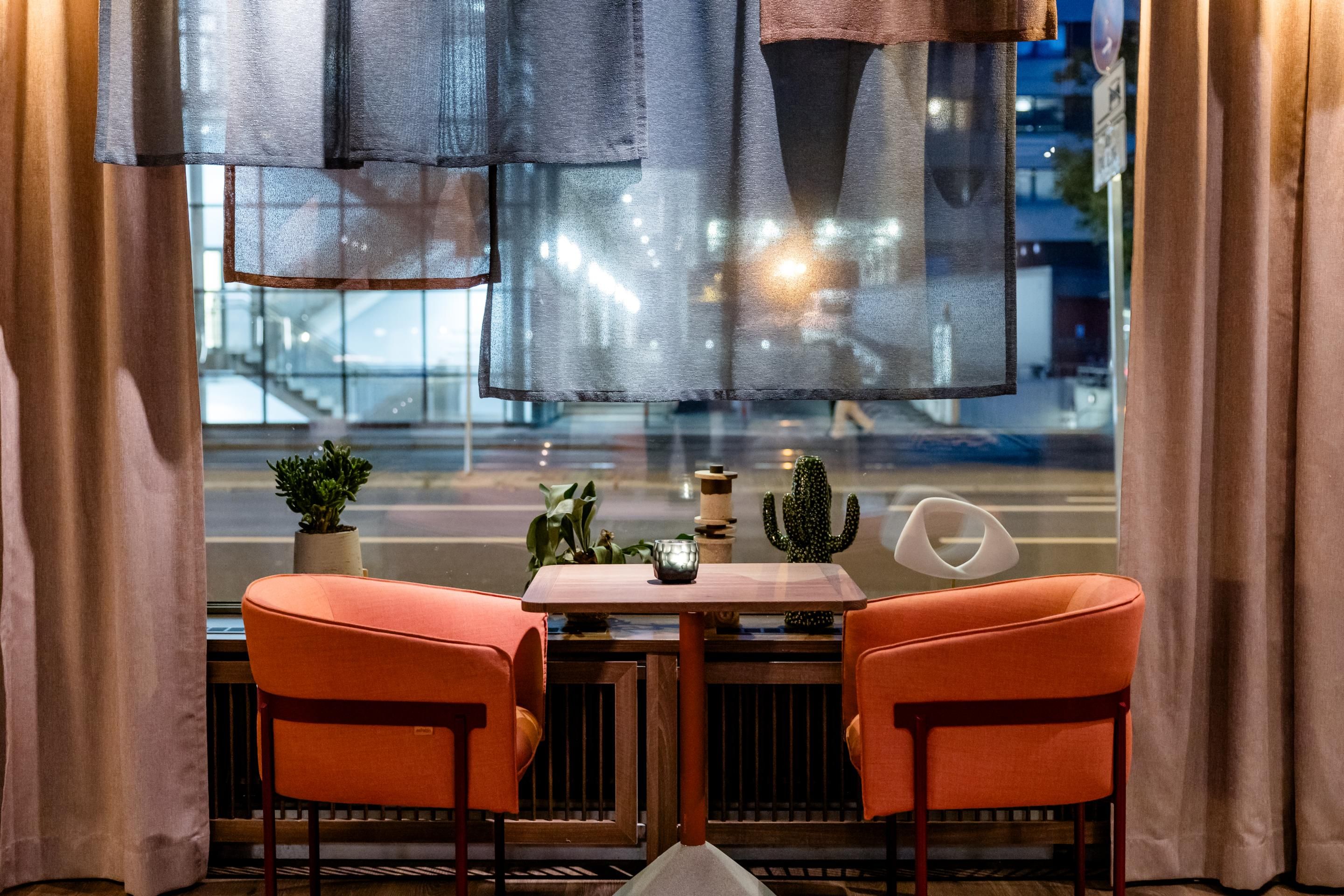 Gemütliche Sitzecke mit zwei orangen Sesseln und kleinem Tisch vor großem Fenster, dekoriert mit Pflanzen. Blick auf beleuchtete Straßenszene im Abendlicht, entspannte Atmosphäre für entspannte Gespräche oder entspanntes Verweilen.