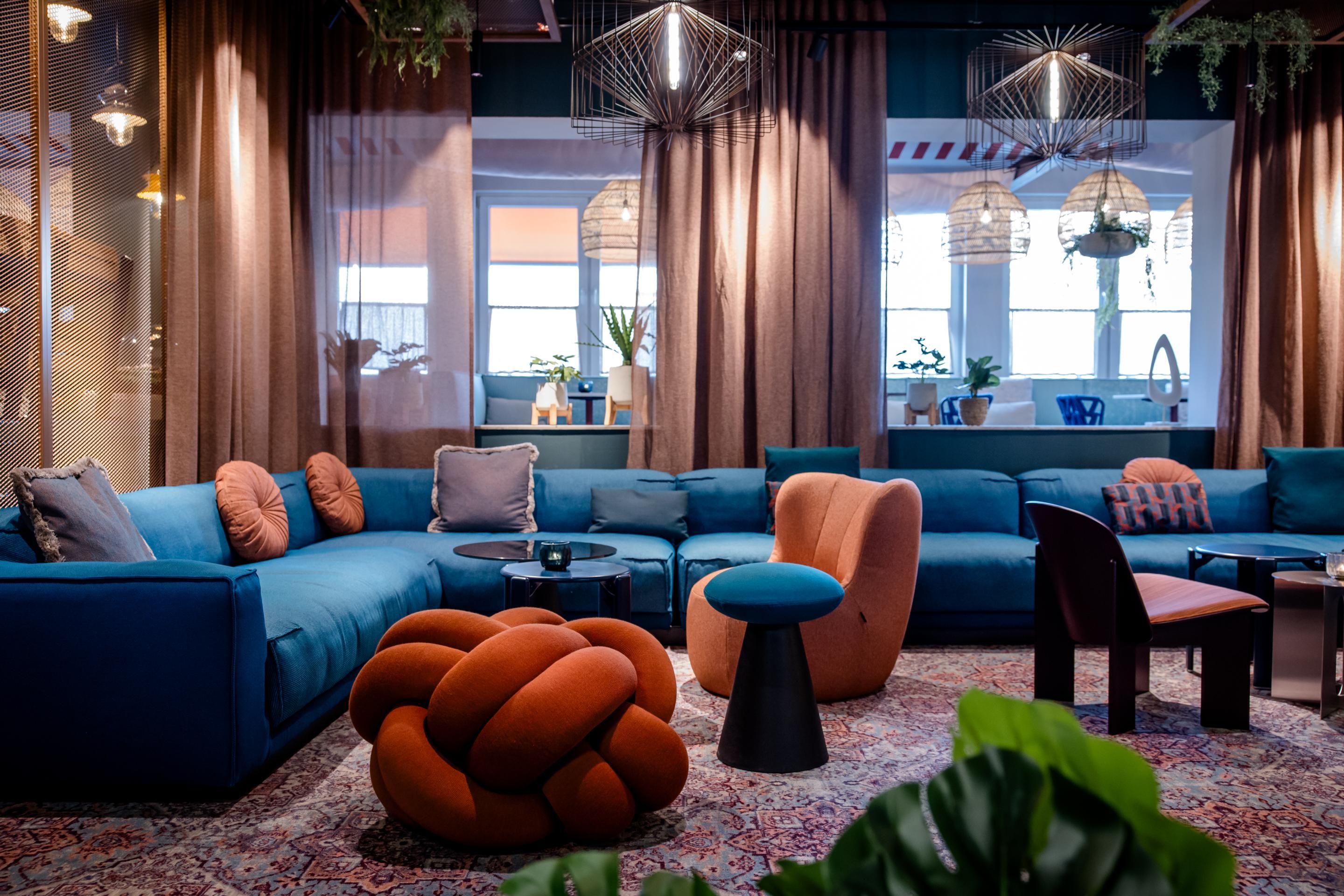 Gemütliche Lounge mit großem blauen Ecksofa, orangen Sesseln und dekorativen Kissen, modernen Hängeleuchten und großen Fenstern, die natürliches Licht hereinlassen. Einladende Atmosphäre für Entspannung und geselliges Beisammensein.