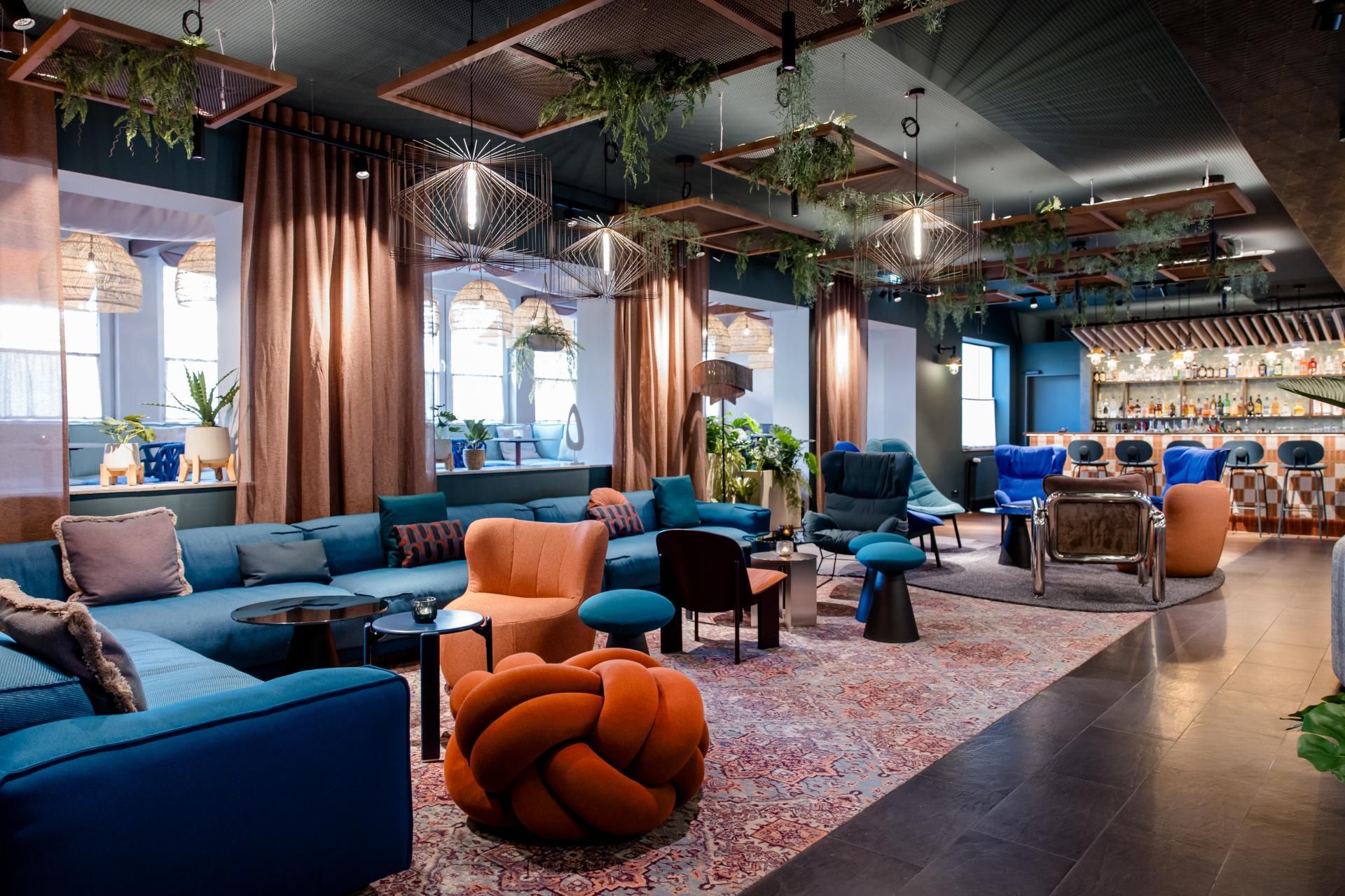 Moderne und gemütliche Hotel-Lounge mit großzügigen Sitzbereichen, bunten Sesseln und Sofas, dekorativen Teppichen und Pflanzen, die eine entspannte Atmosphäre für Gäste schaffen. Sanfte Beleuchtung und nachhaltige Design-Elemente sorgen für Wohlbefinden und stilvolles Ambiente.
