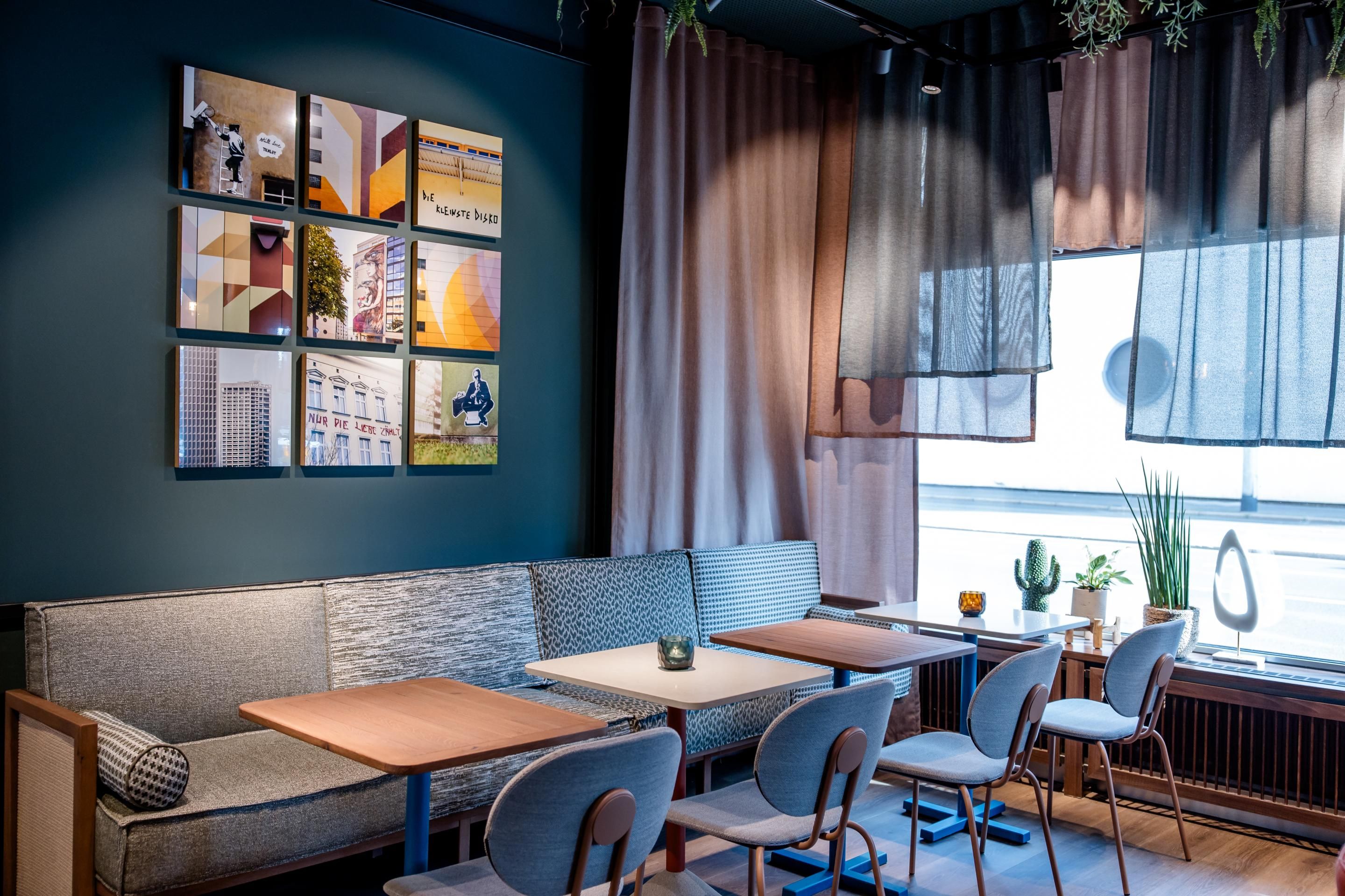 Gemütlicher Lounge-Bereich mit modernem Sofa, kleinen Holztischen und gepolsterten Stühlen. Große Fenster mit halbtransparenten Vorhängen lassen Tageslicht herein. An der Wand dekorative Bilder mit urbanen Motiven schaffen eine entspannte Atmosphäre. Ideal für entspannte Pausen oder geselliges Beisammensein.