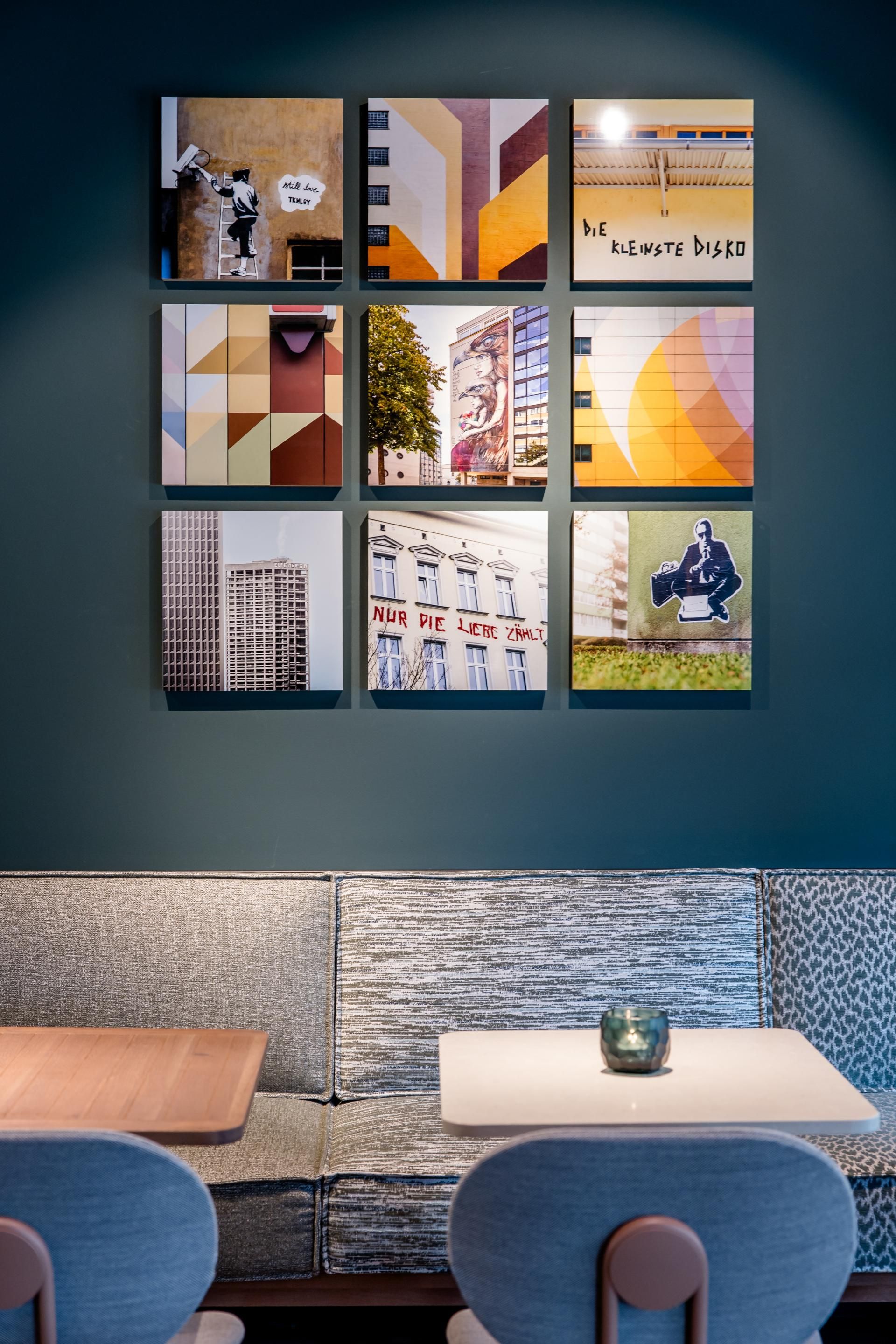 Neun quadratische Bilder mit urbaner Kunst und grafischen Mustern hängen über einer modernen Lounge-Ecke mit einer grauen Polsterbank und zwei Stühlen, die eine gemütliche Atmosphäre schaffen.