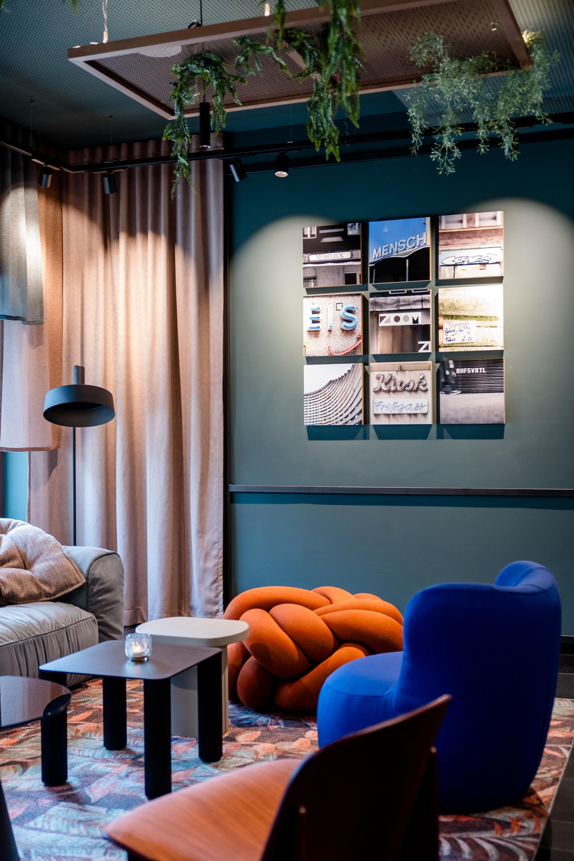 Gemütliche Lounge mit modernem Mobiliar: Bequemer grauer Sessel, knallblauer Schaukelstuhl, orangefarbener Knoten-Pouf, schwarzer Couchtisch und grüne Hängepflanzen, die eine entspannte Atmosphäre schaffen. An der Wand dekorative Bilder mit Schriftzügen, weiche Vorhänge sorgen für gedämpftes Licht.