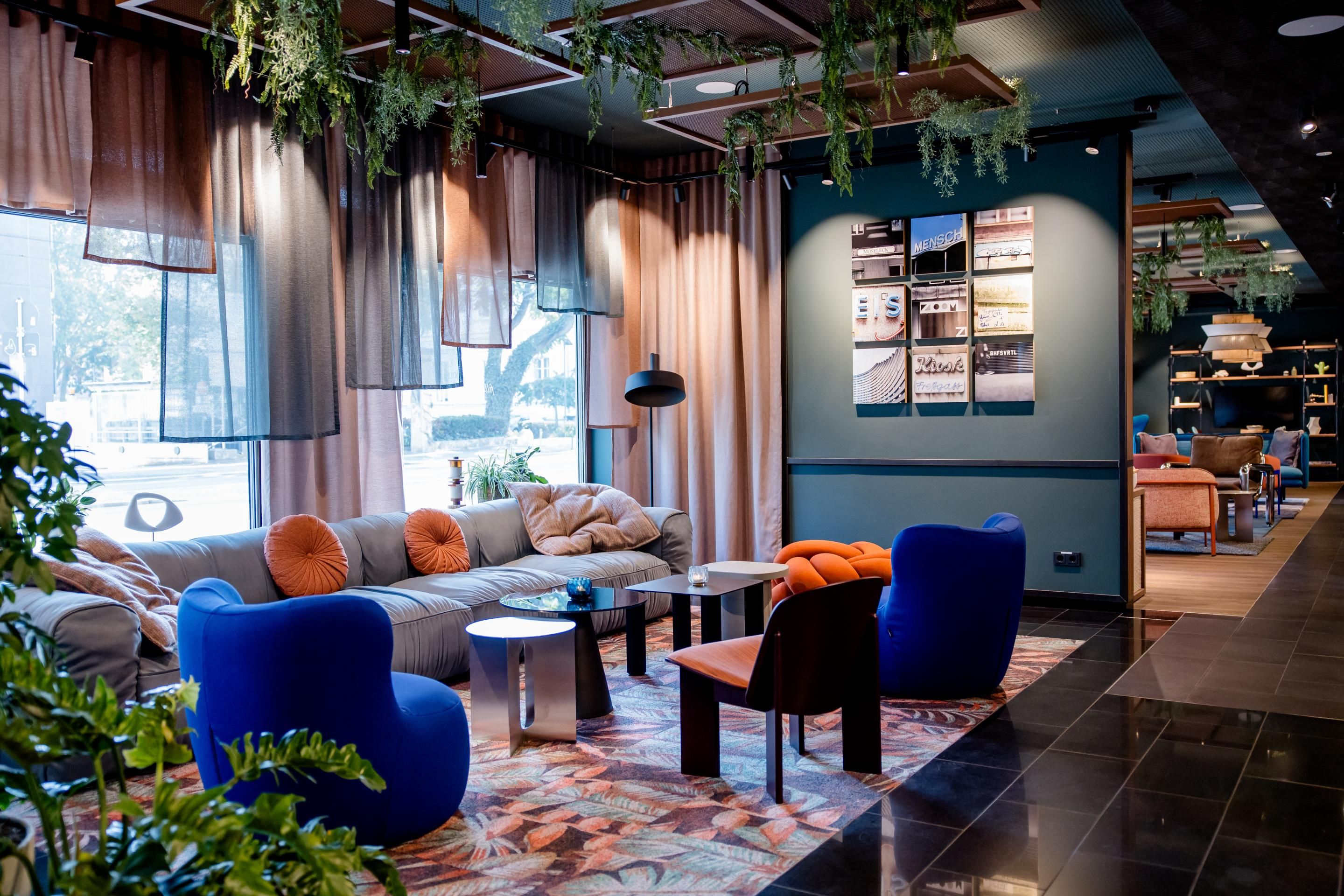 Eleganter, gemütlicher Lounge-Bereich mit Sofas, bunten Sesseln, Teppich und großen Fenstern, umgeben von Pflanzen und warmem Licht, ideal für entspannte Gespräche oder ruhige Momente.