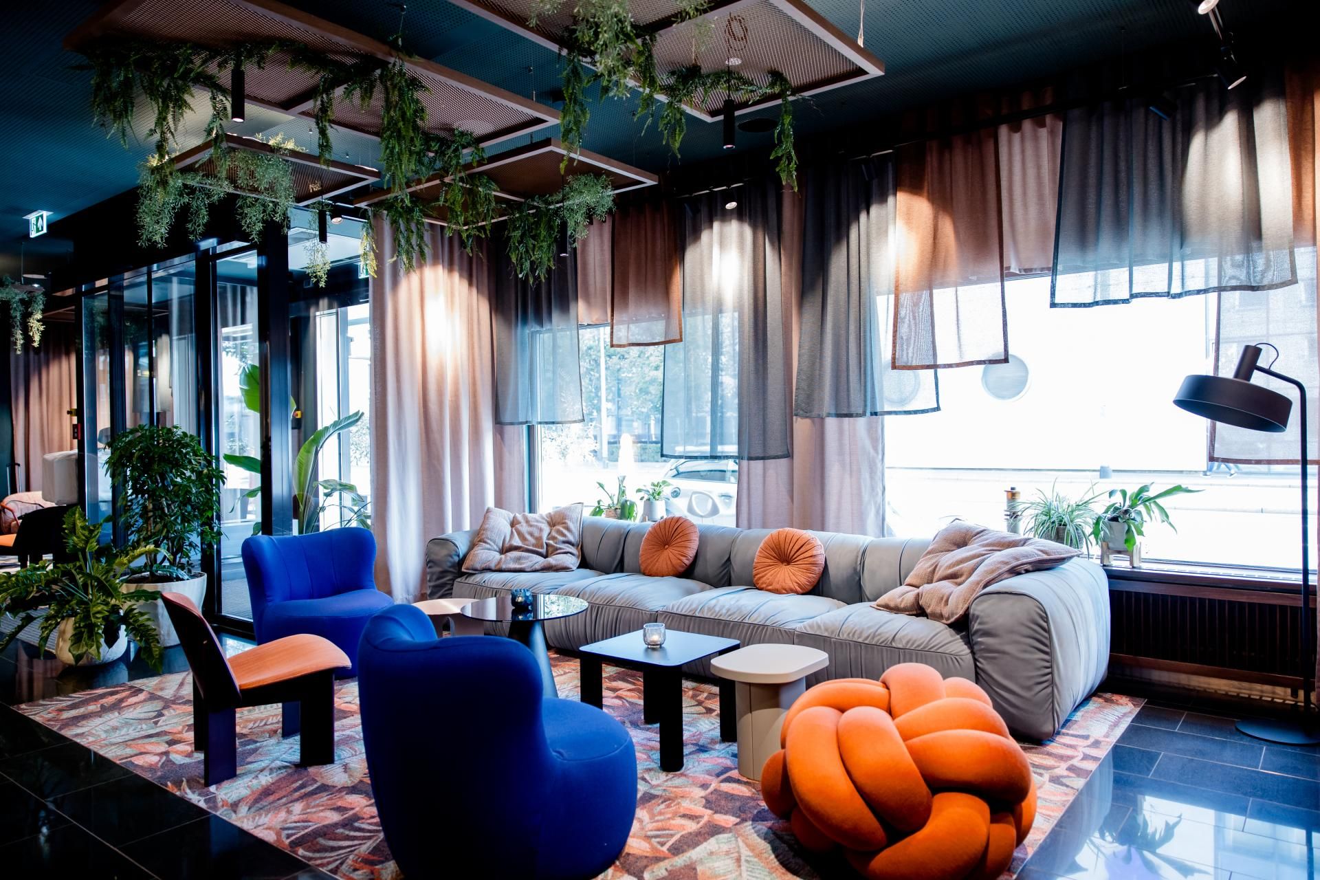 Gemütliche Lounge mit großem grauem Sofa, bunten Sesseln und Pflanzen, sanfte Beleuchtung und Vorhänge schaffen eine einladende Atmosphäre zum Entspannen und Verweilen.