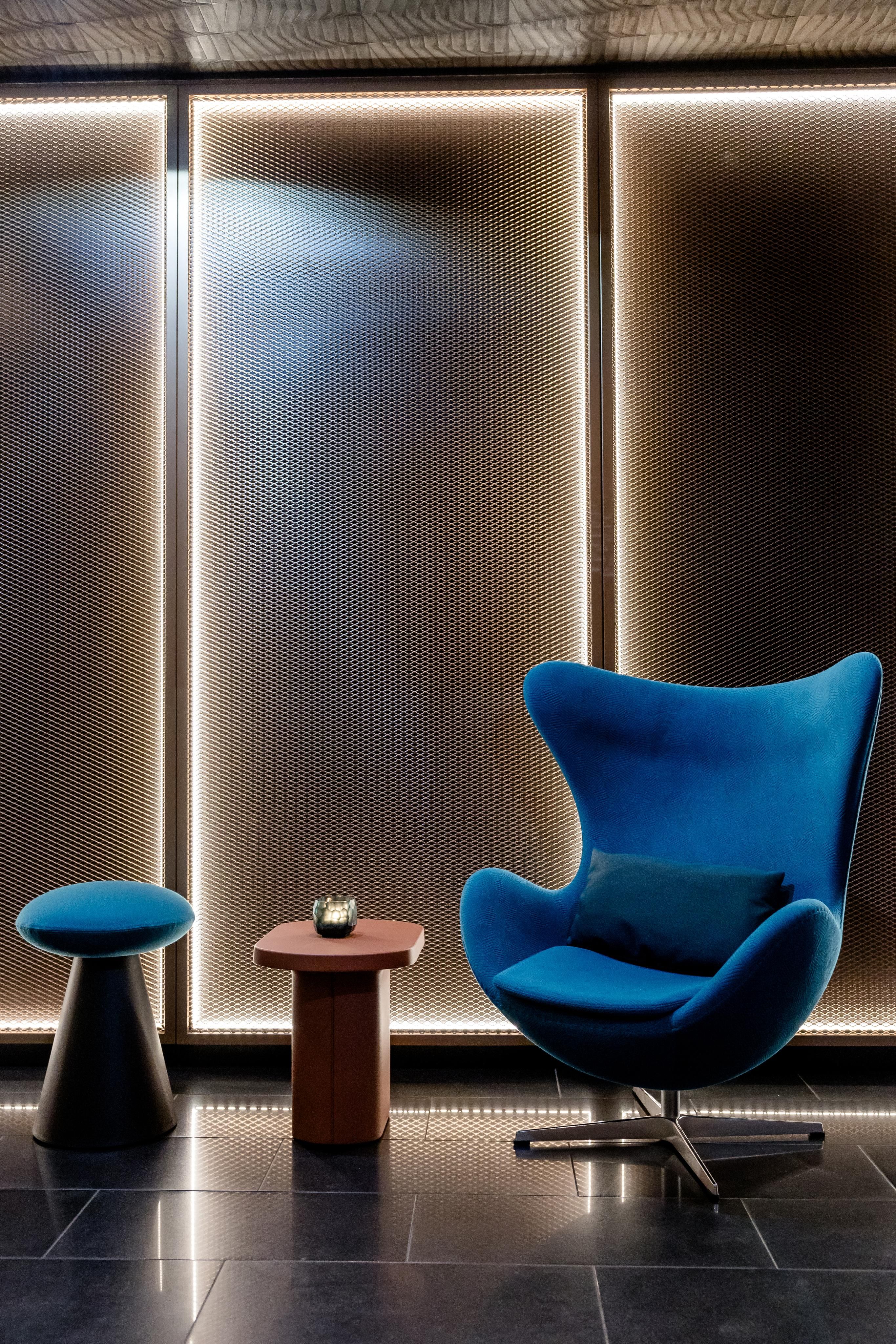 Moderne Leseecke mit blauem Designer-Sessel, passendem Hocker und kleinem Holztisch vor samtener, indirekt beleuchteter Wand, ideal für Entspannung und ruhige Momente im Hotelambiente.