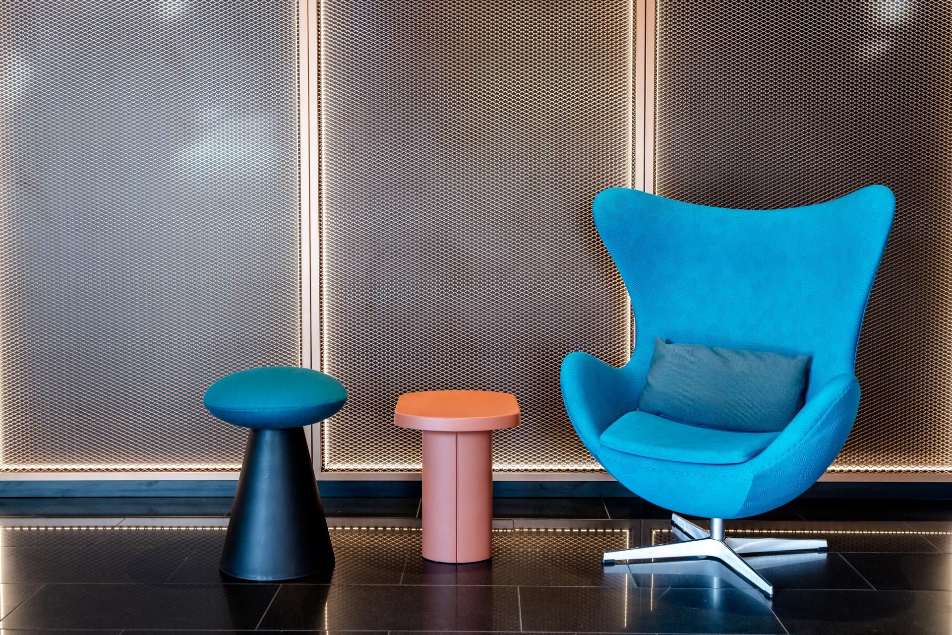Moderner, gemütlicher Sitzbereich mit einem auffällig blauen Sessel und zwei kleinen Beistelltischen vor einer beleuchteten, metallischen Wand, ideal zum Entspannen oder für kurze Gespräche.