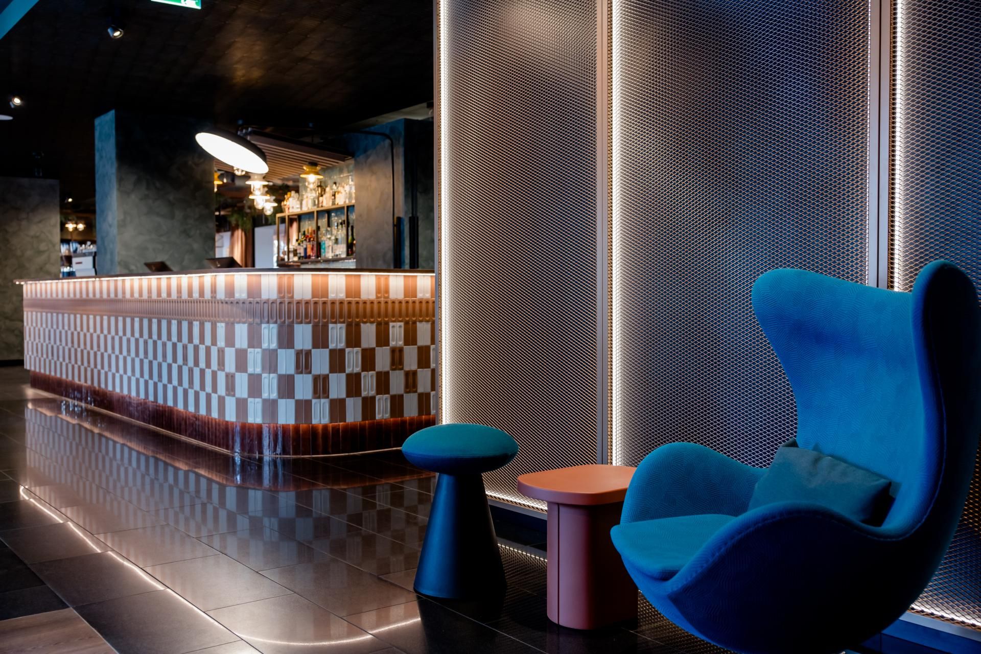 Moderne Hotelbar mit stilvoll beleuchtetem Tresen und gemütlicher Sitzecke mit blauem Design-Sessel und kleinen Tischen, die eine entspannte Atmosphäre für Gäste schaffen.