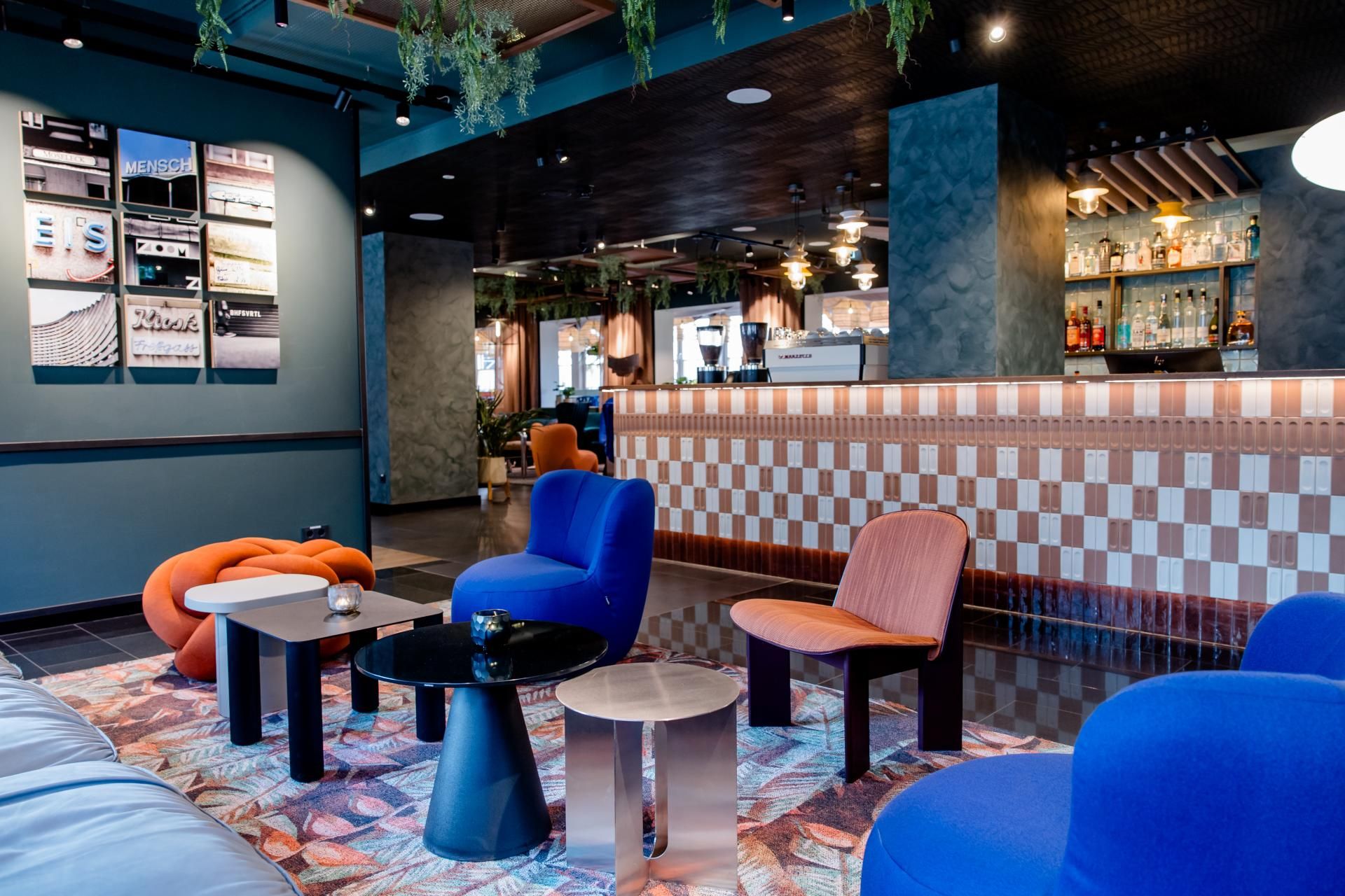 Moderne Hotelbar mit gemütlichen, farbenfrohen Sesseln und Tischen, dekorativen Hängepflanzen und stilvoller Beleuchtung, die eine einladende Atmosphäre zum Entspannen und Verweilen schafft.