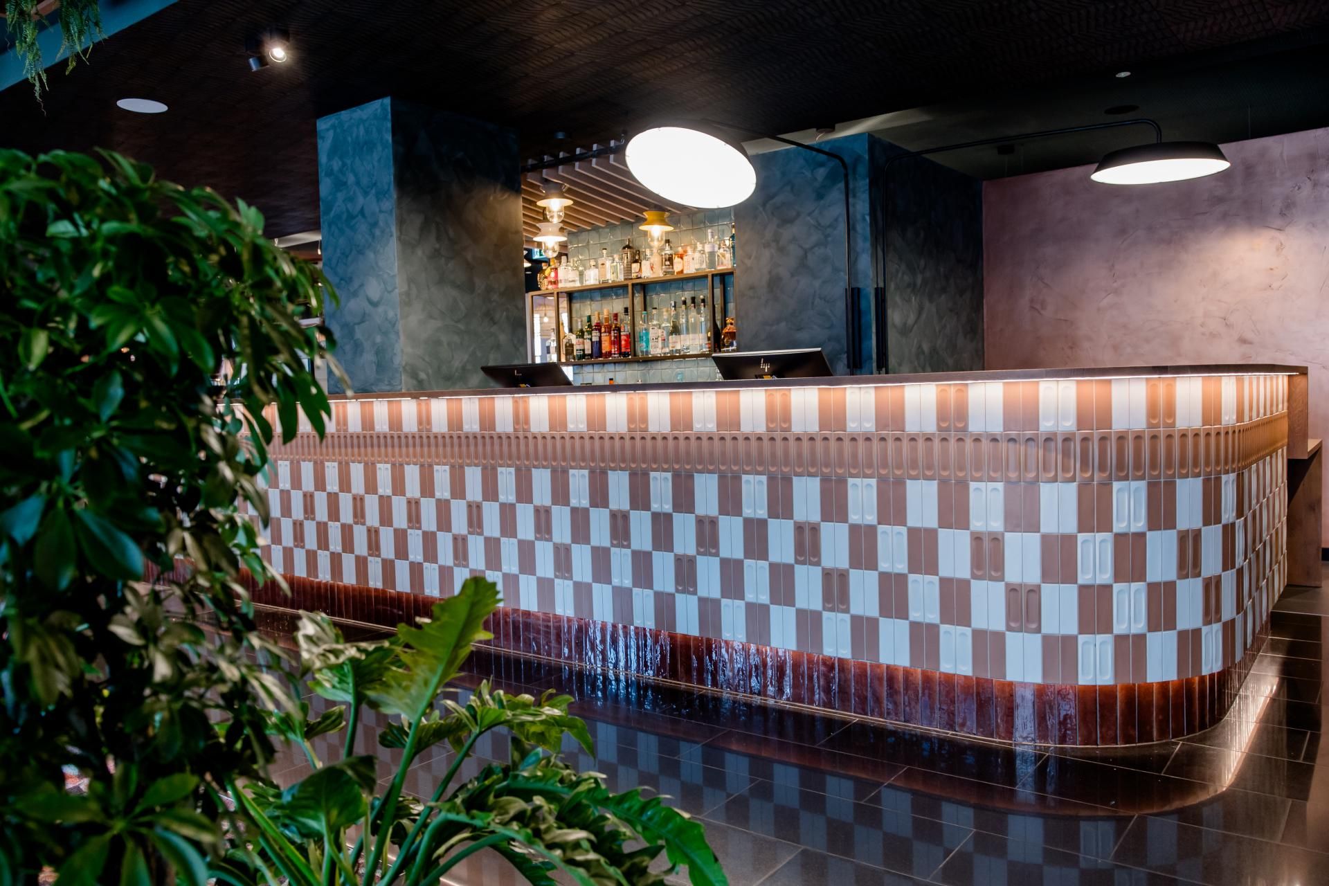 Moderne Hotelbar mit stilvollem, geometrischem Fliesenmuster an der Theke, warmem Licht und grünen Pflanzen im Vordergrund, die eine einladende und entspannte Atmosphäre schaffen.