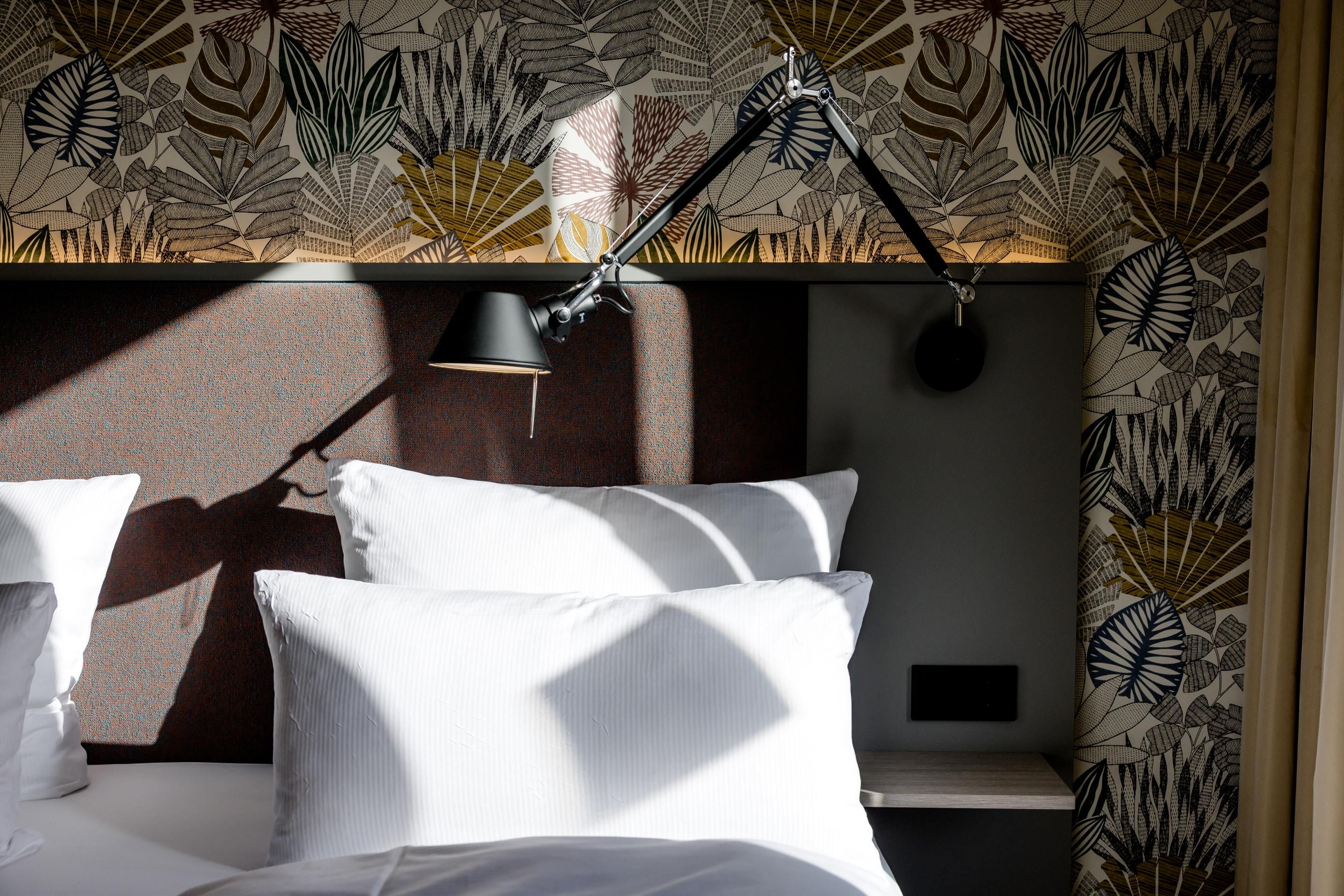 Helles, gemütliches Hotelzimmer mit weißen Kissen und Bettwäsche. Moderne Leselampe am Kopfteil, das mit einem stilvollen, botanisch gemusterten Tapetenhintergrund dekoriert ist. Sanftes Tageslicht beleuchtet die ruhige Schlafumgebung.