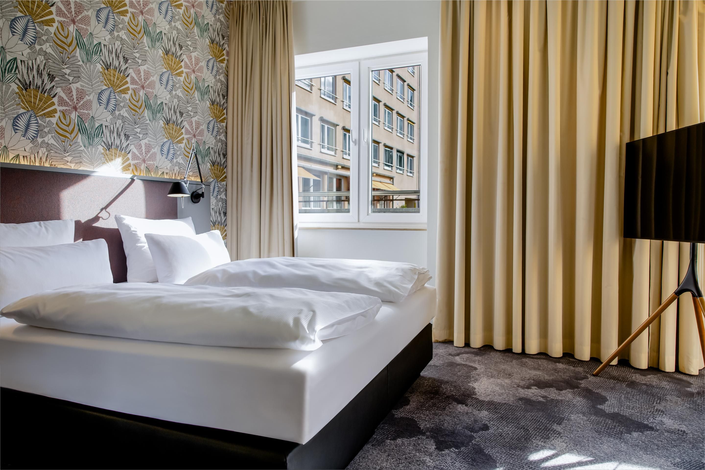 Helles Hotelzimmer mit großem, gemütlichem Doppelbett, floraler Akzentwand, großem Fenster mit Stadtblick und beigen Vorhängen, ruhig und einladend gestaltet.