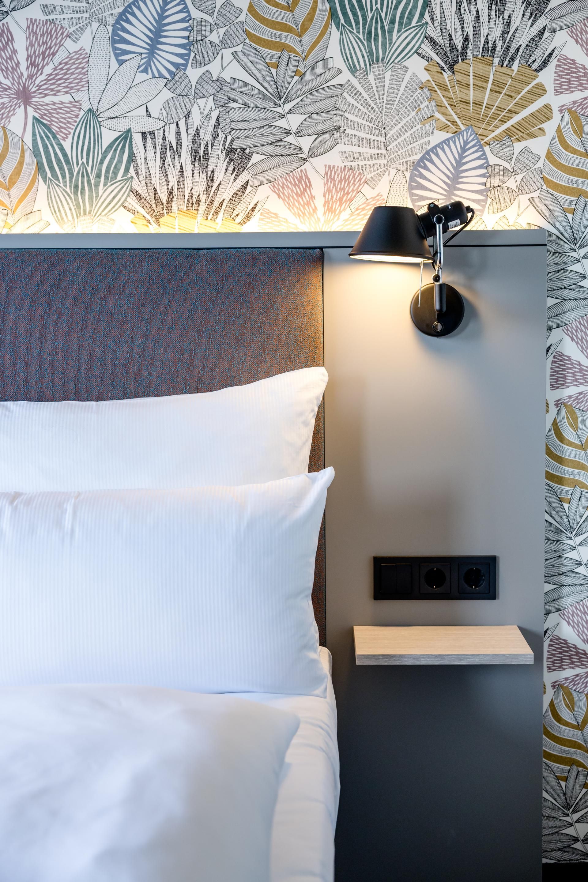 Bett mit weißer Bettwäsche, grauem gepolstertem Kopfteil und Nachttisch mit moderner Wandlampe, umgeben von Tapete mit farbenfrohem Blattmuster.