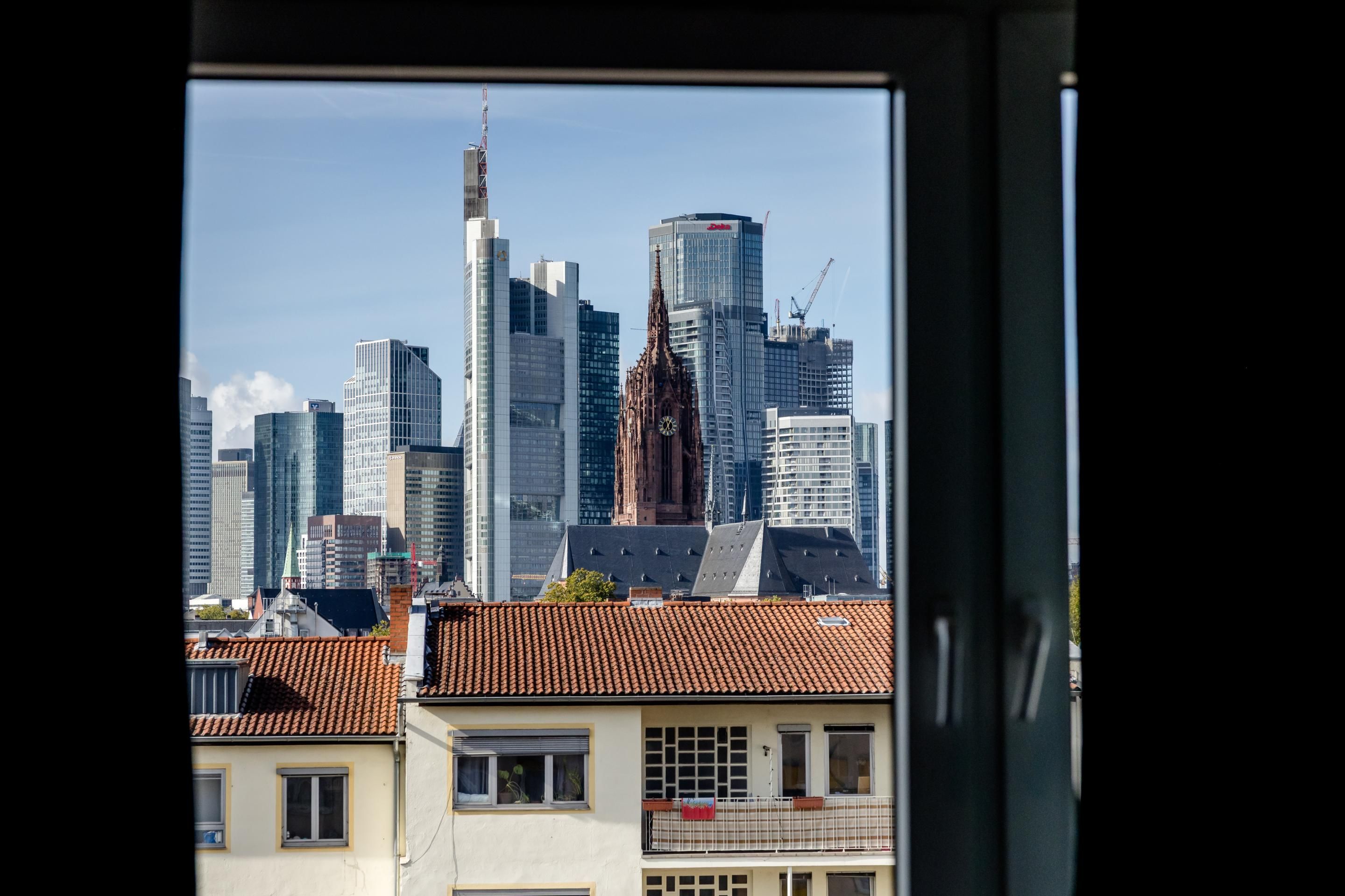 Blick aus einem Fenster auf die Frankfurter Skyline mit modernen Wolkenkratzern und der historischen Paulskirche, umgeben von Wohngebäuden mit roten Dächern.
