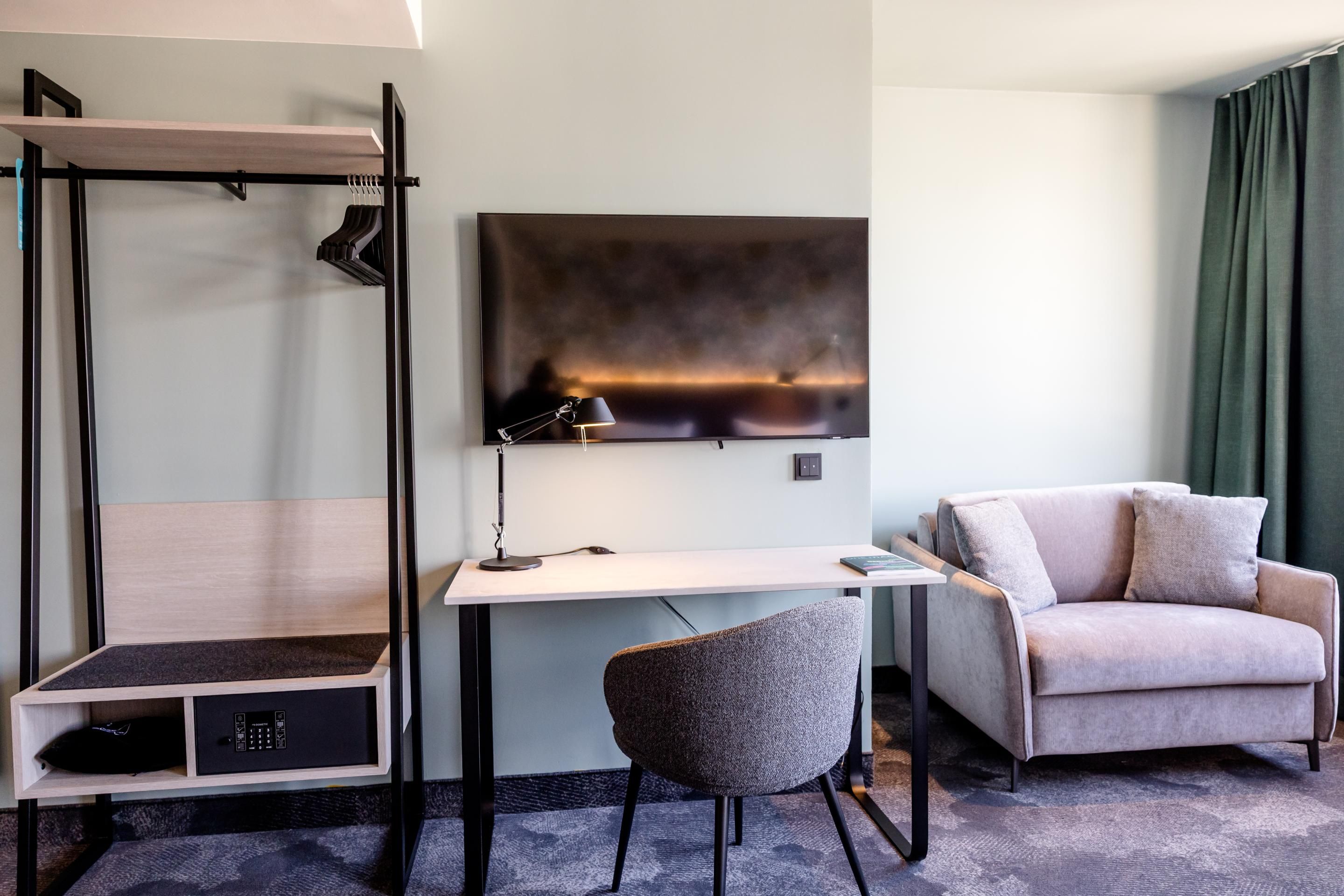 Modern eingerichteter Hotelzimmerbereich mit Schreibtisch, Stuhl, Wand-TV, Sofa und offenem Kleiderständer, in ruhigen, neutralen Farben für entspannte Arbeits- und Aufenthaltsmöglichkeiten.