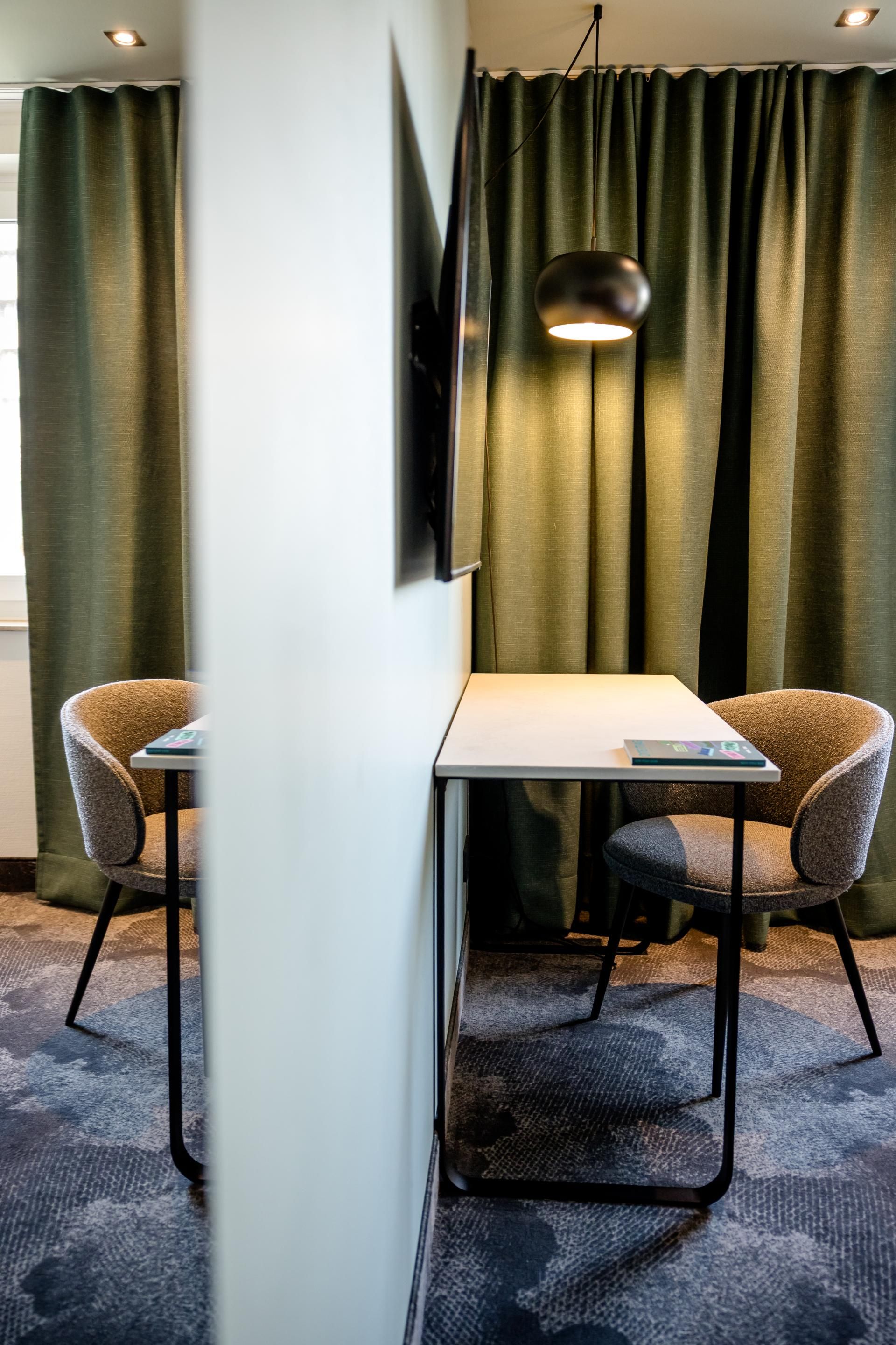 Kleiner Arbeitsplatz mit Tisch, Stuhl und Hängeleuchte vor dunklen Vorhängen in einem ruhigen Hotelzimmer, ideal für konzentriertes Arbeiten oder Lesen.