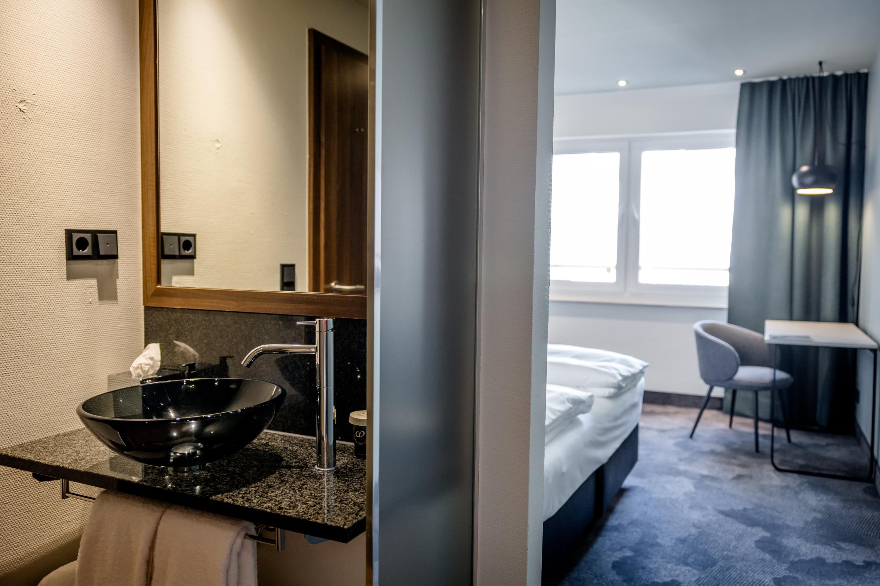 Blick in ein modernes Hotelzimmer mit hellen Wänden, komfortablem Bett, Schreibtisch und Stuhl. Separater Waschbereich mit schwarzem Waschbecken und großem Spiegel. Tageslicht durch großes Fenster, ruhige und einladende Atmosphäre für Erholung und Arbeit.