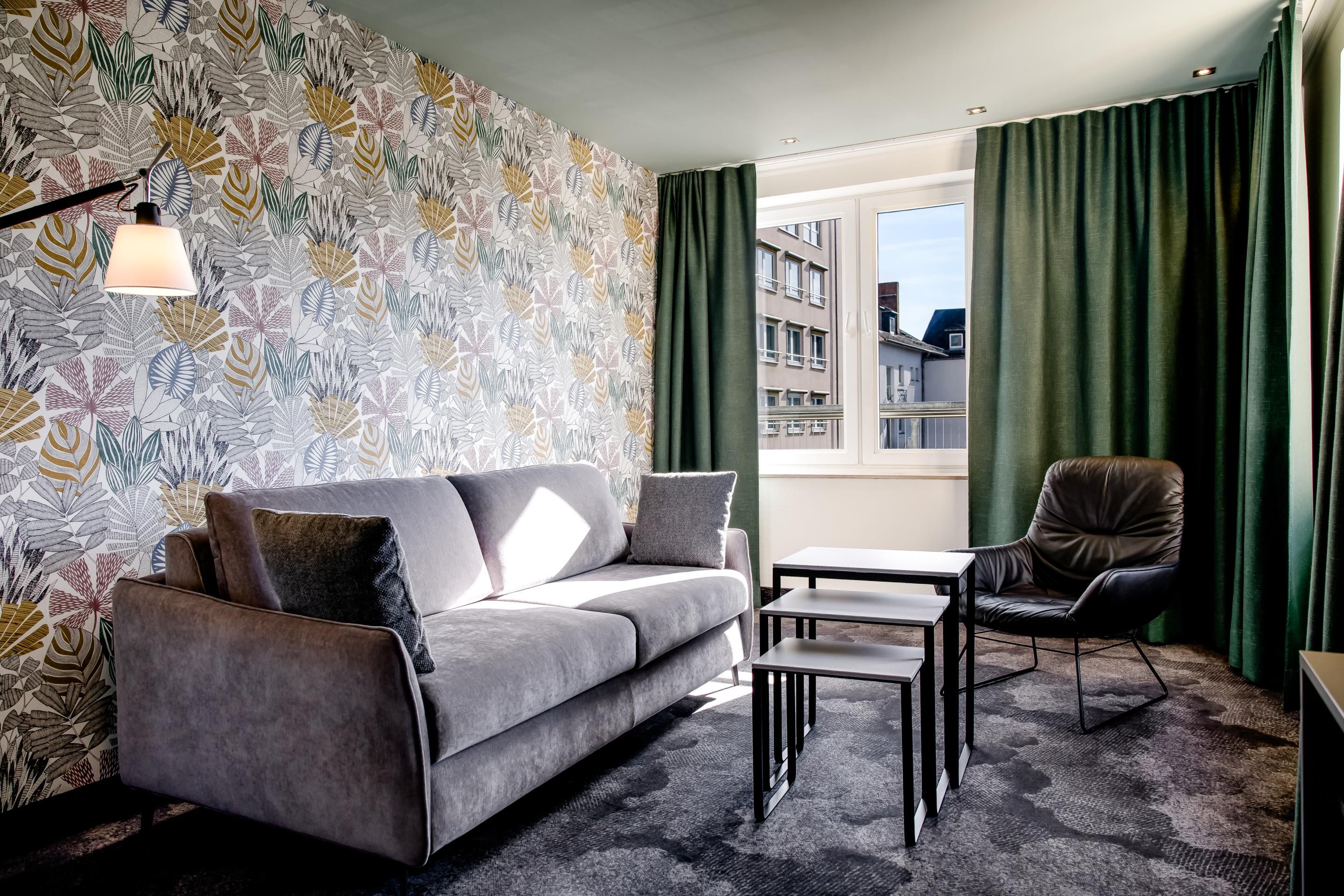 Modern eingerichtetes Wohnzimmer mit grauem Sofa, schwarzem Sessel, Tapeten mit Blattmuster, drei ineinander passenden Couchtischen und großem Fenster mit Blick auf umliegende Gebäude.