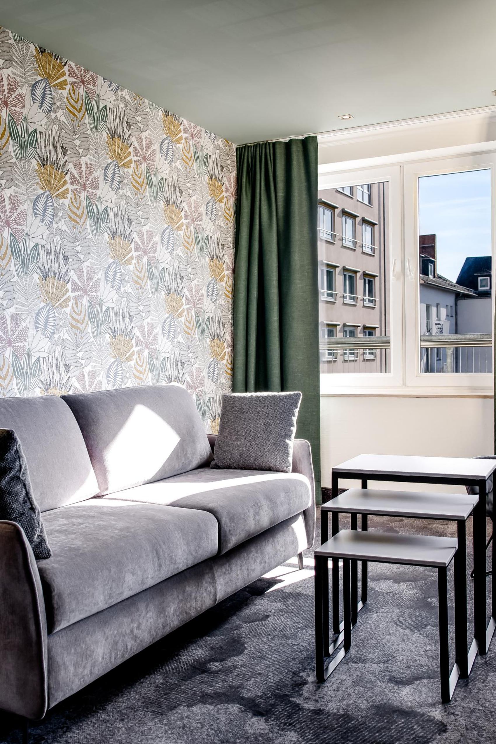Modern eingerichtetes Wohnzimmer mit grauem Sofa, schwarzem Sessel, Tapeten mit Blattmuster, drei ineinander passenden Couchtischen und großem Fenster mit Blick auf umliegende Gebäude.