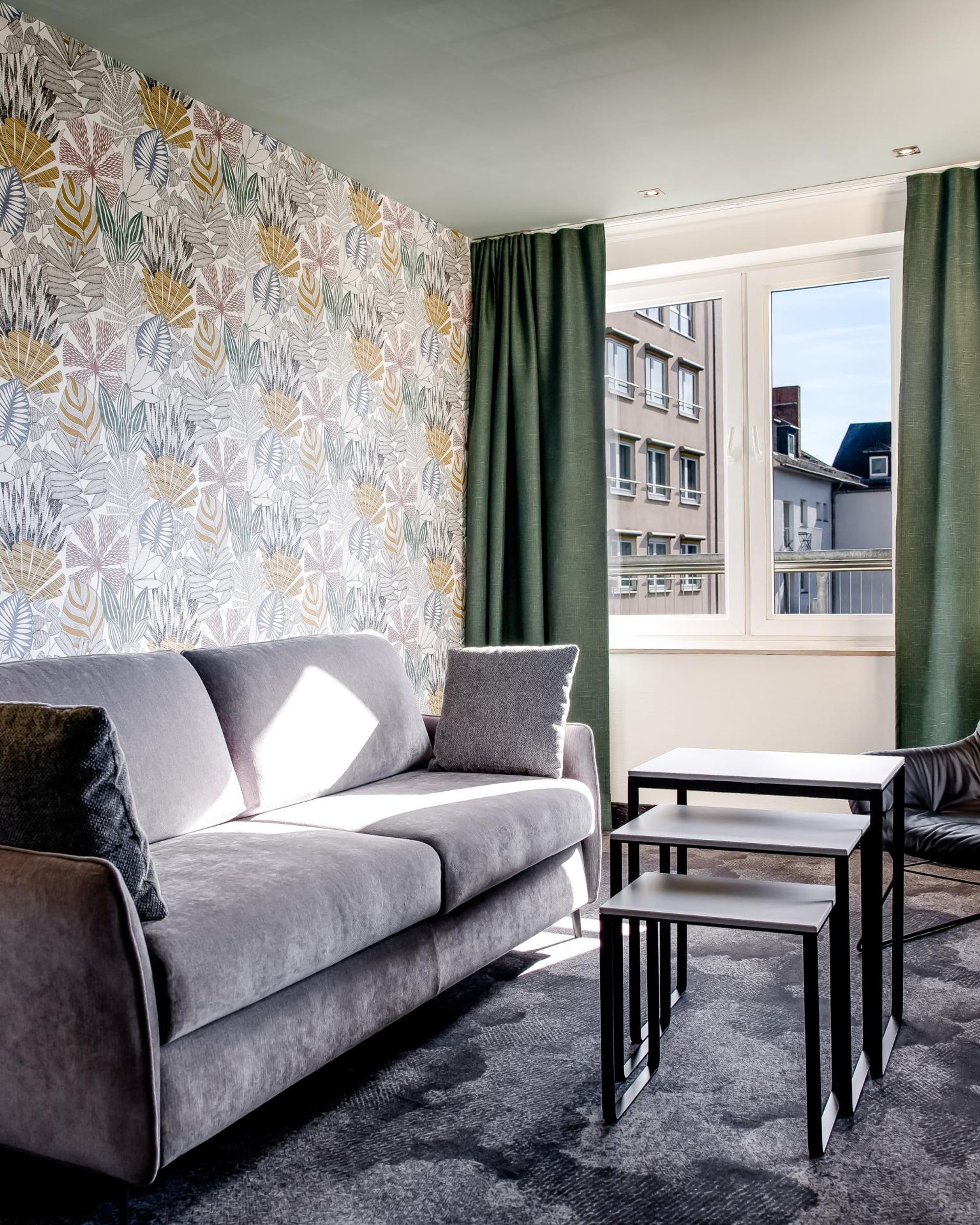 Modern eingerichtetes Wohnzimmer mit grauem Sofa, schwarzem Sessel, Tapeten mit Blattmuster, drei ineinander passenden Couchtischen und großem Fenster mit Blick auf umliegende Gebäude.