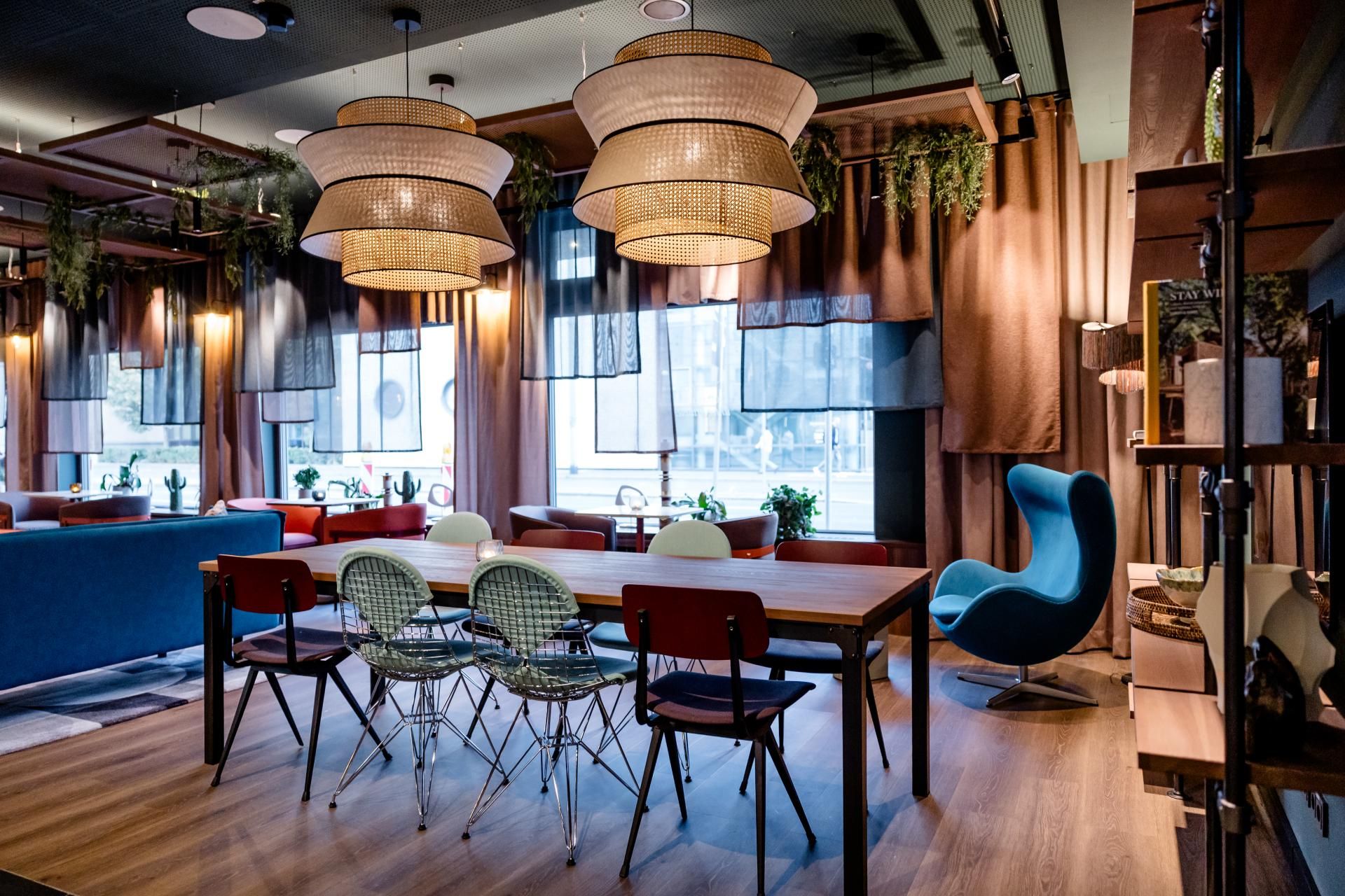Moderne, gemütliche Lounge mit großem Holztisch, verschiedenen Stühlen und entspannter Beleuchtung durch große, geflochtene Lampenschirme in einem stilvollen, einladenden Innenraum.