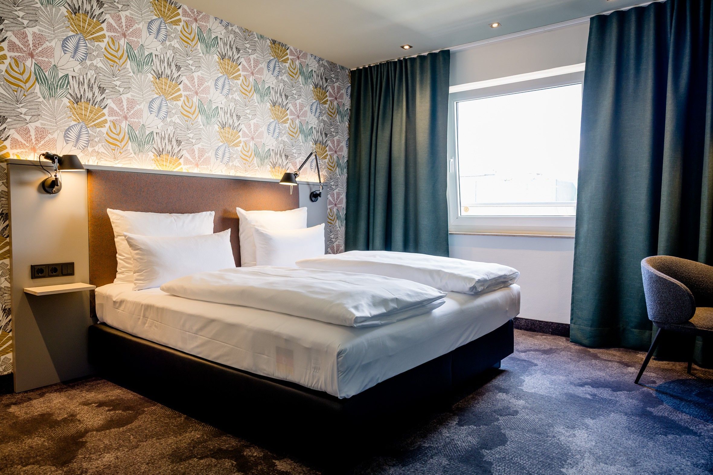 Modern eingerichtetes Hotelzimmer mit großem Doppelbett, floral gemusterter Akzentwand und großem Fenster mit dunklen Vorhängen, das eine helle und einladende Atmosphäre schafft.