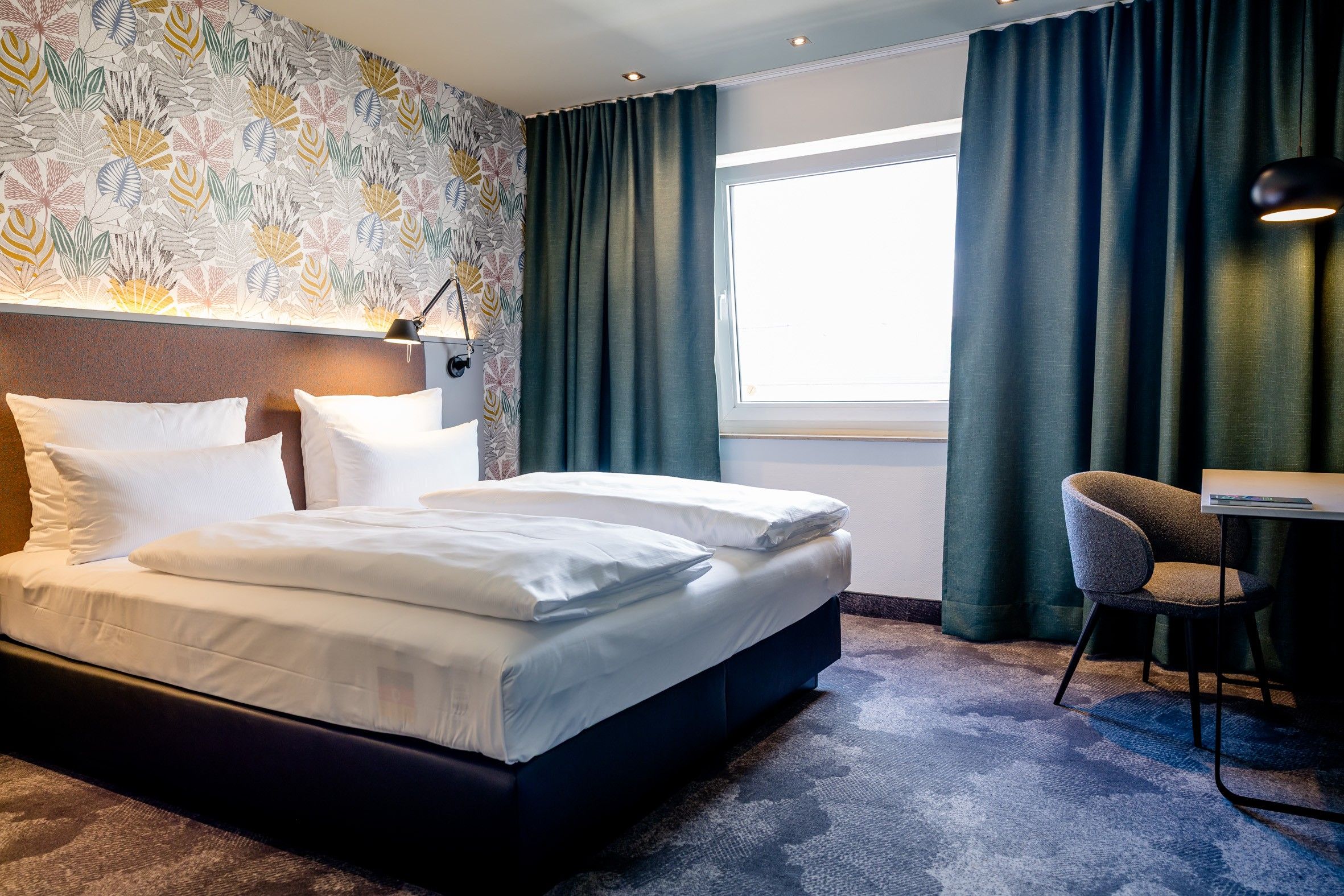 Modernes Hotelzimmer mit großem Doppelbett, dekorativer Tapete, dunkelgrünen Vorhängen und einem kleinen Sitzbereich mit Stuhl und Schreibtisch. Helle und gemütliche Atmosphäre, ideal für Erholung und Arbeit.
