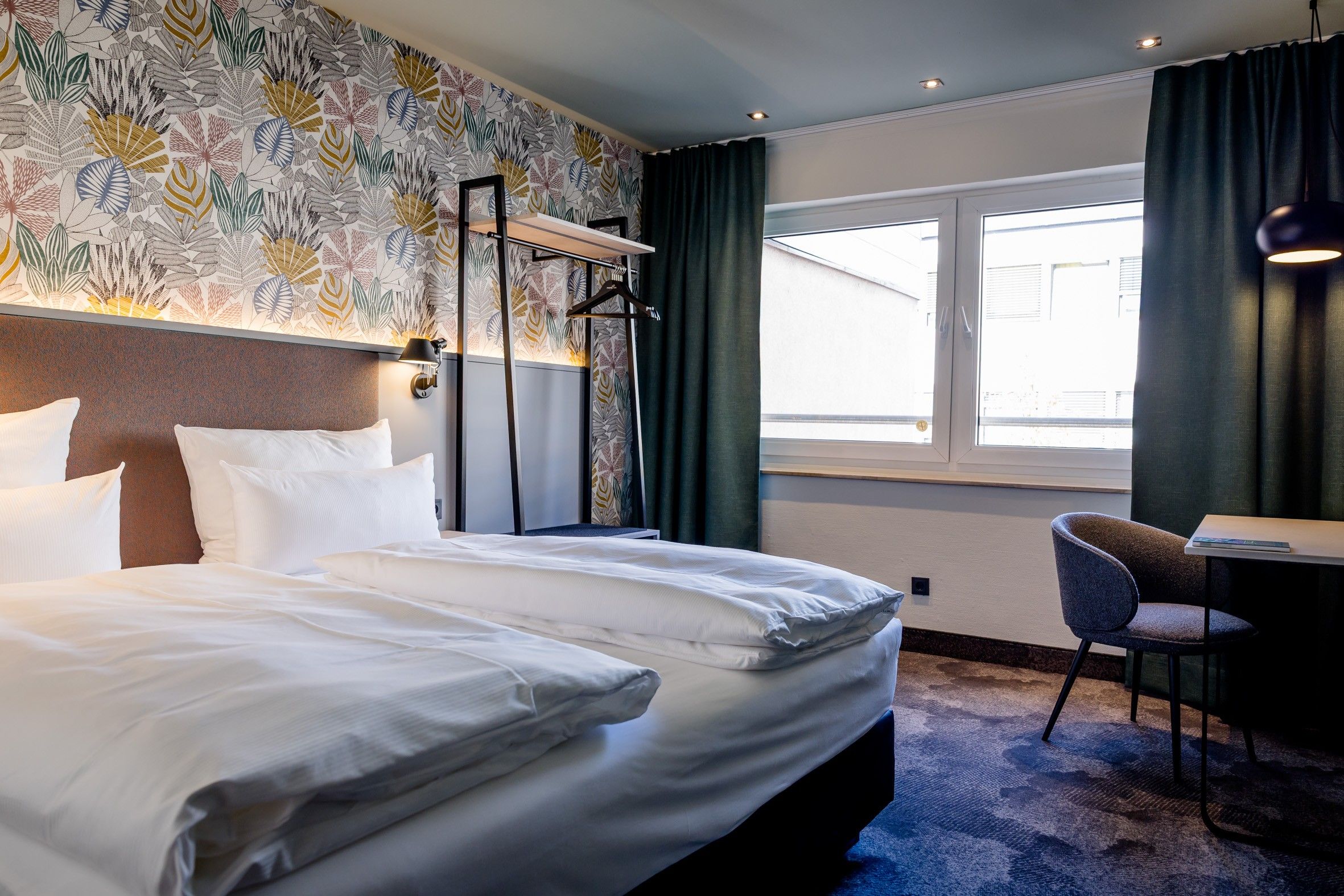 Modernes Hotelzimmer mit zwei Einzelbetten, stilvoller Tapete, großem Fenster und gemütlichem Arbeitsplatz, ideal für erholsamen Schlaf und produktives Arbeiten.