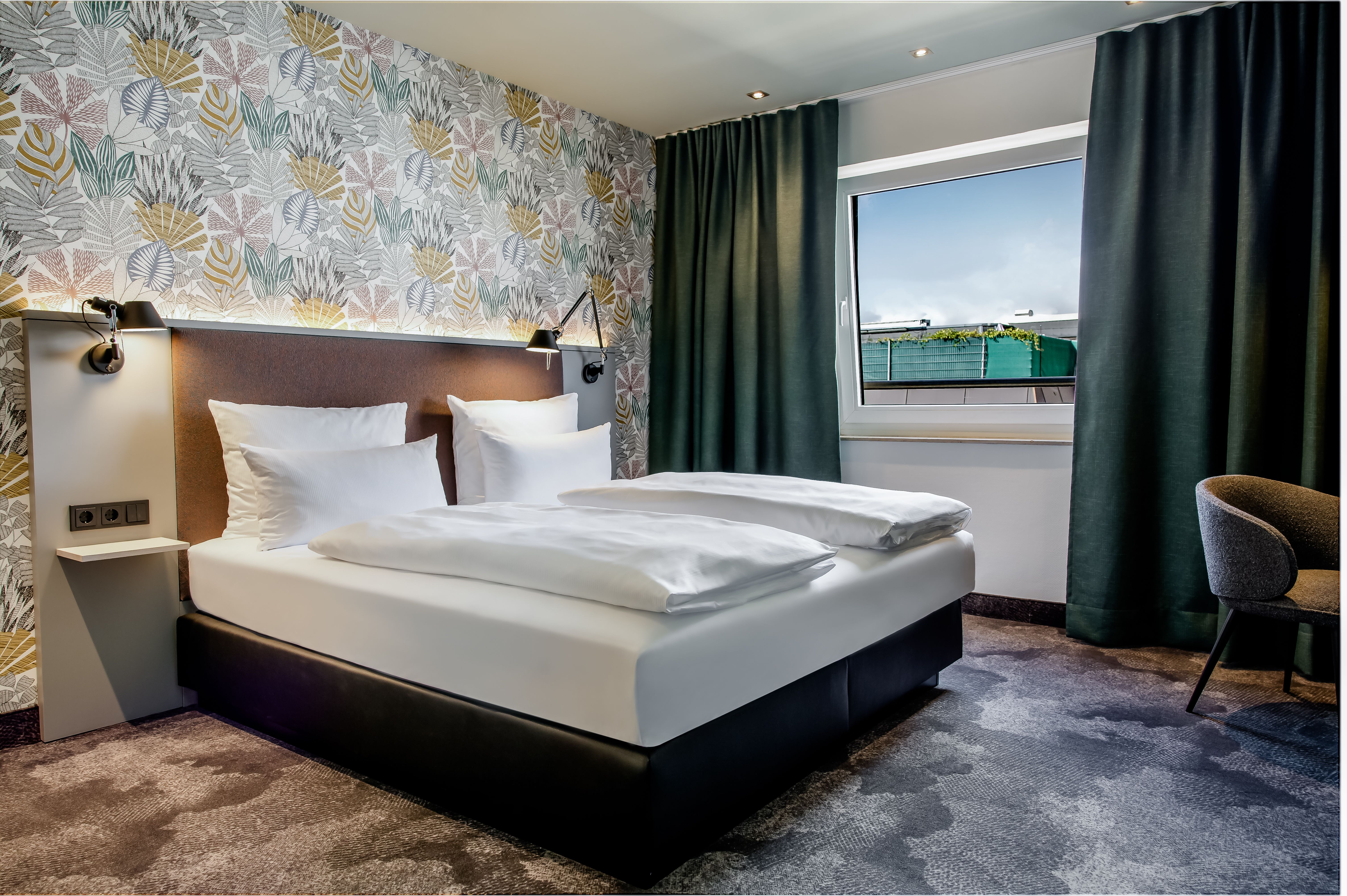Modern eingerichtetes Hotelzimmer mit Doppelbett, gemütlicher Bettwäsche, dekorativer Wand mit Pflanzenmuster, großem Fenster mit Verdunkelungsvorhängen und einem Stuhl, das eine ruhige und entspannte Atmosphäre vermittelt.