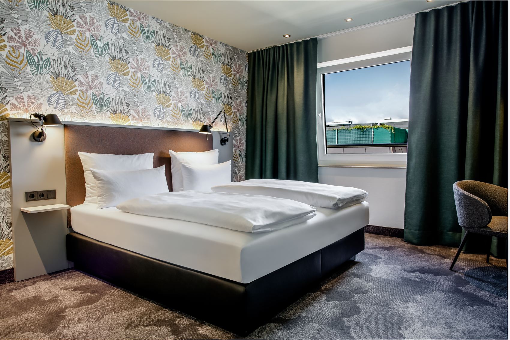Modern eingerichtetes Hotelzimmer mit Doppelbett, gemütlicher Bettwäsche, dekorativer Wand mit Pflanzenmuster, großem Fenster mit Verdunkelungsvorhängen und einem Stuhl, das eine ruhige und entspannte Atmosphäre vermittelt.