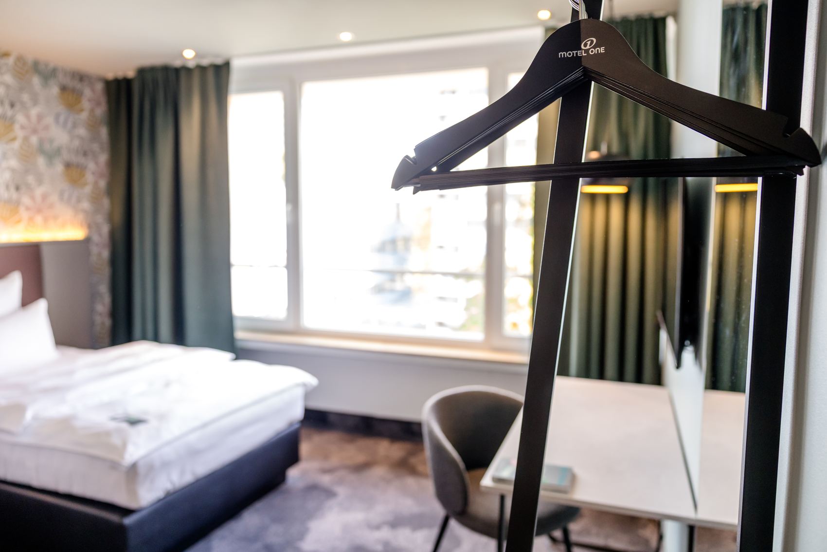 Modern eingerichtetes Hotelzimmer mit großem Fenster, bequemen Doppelbett, Schreibtisch und Stuhl, ergänzt durch dunkle Vorhänge für angenehme Verdunkelung.