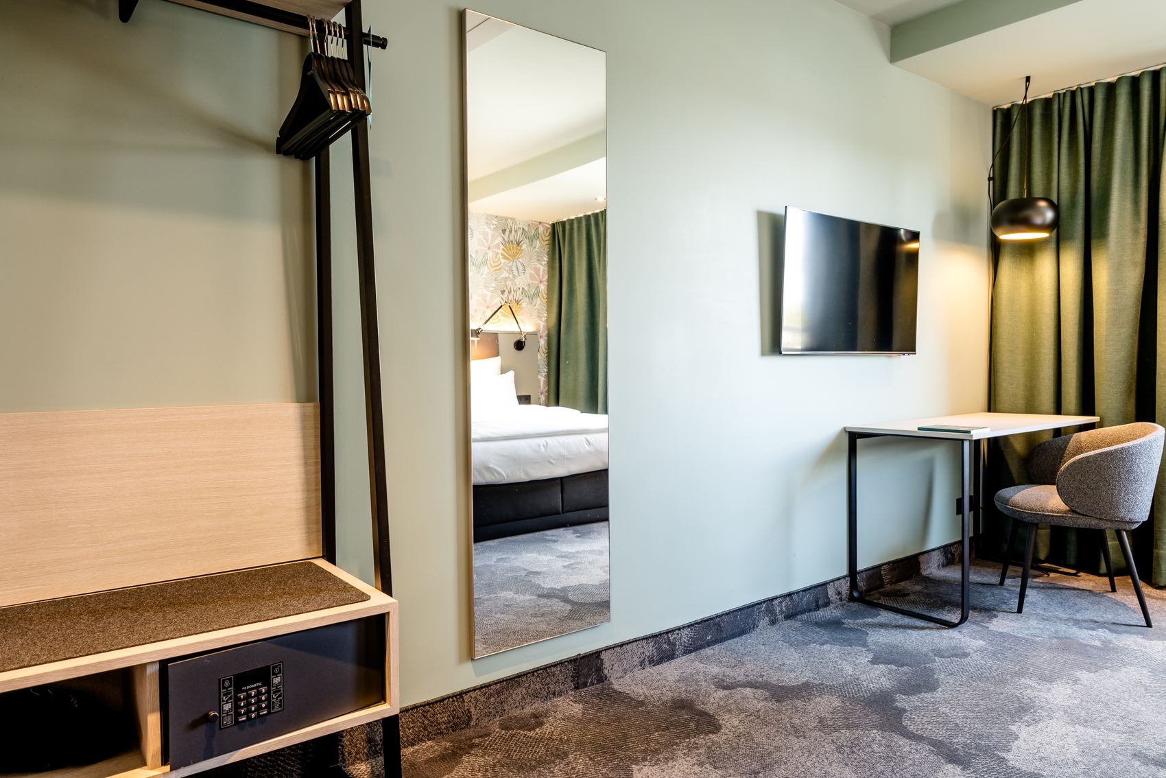 Modern eingerichtetes Hotelzimmer mit Sitzbereich, großem Spiegel, Flat-TV und Garderobe mit Safe. Gemütliche Atmosphäre durch sanfte Beleuchtung und Teppichboden. Teilweise sichtbares Bett im Hintergrund.