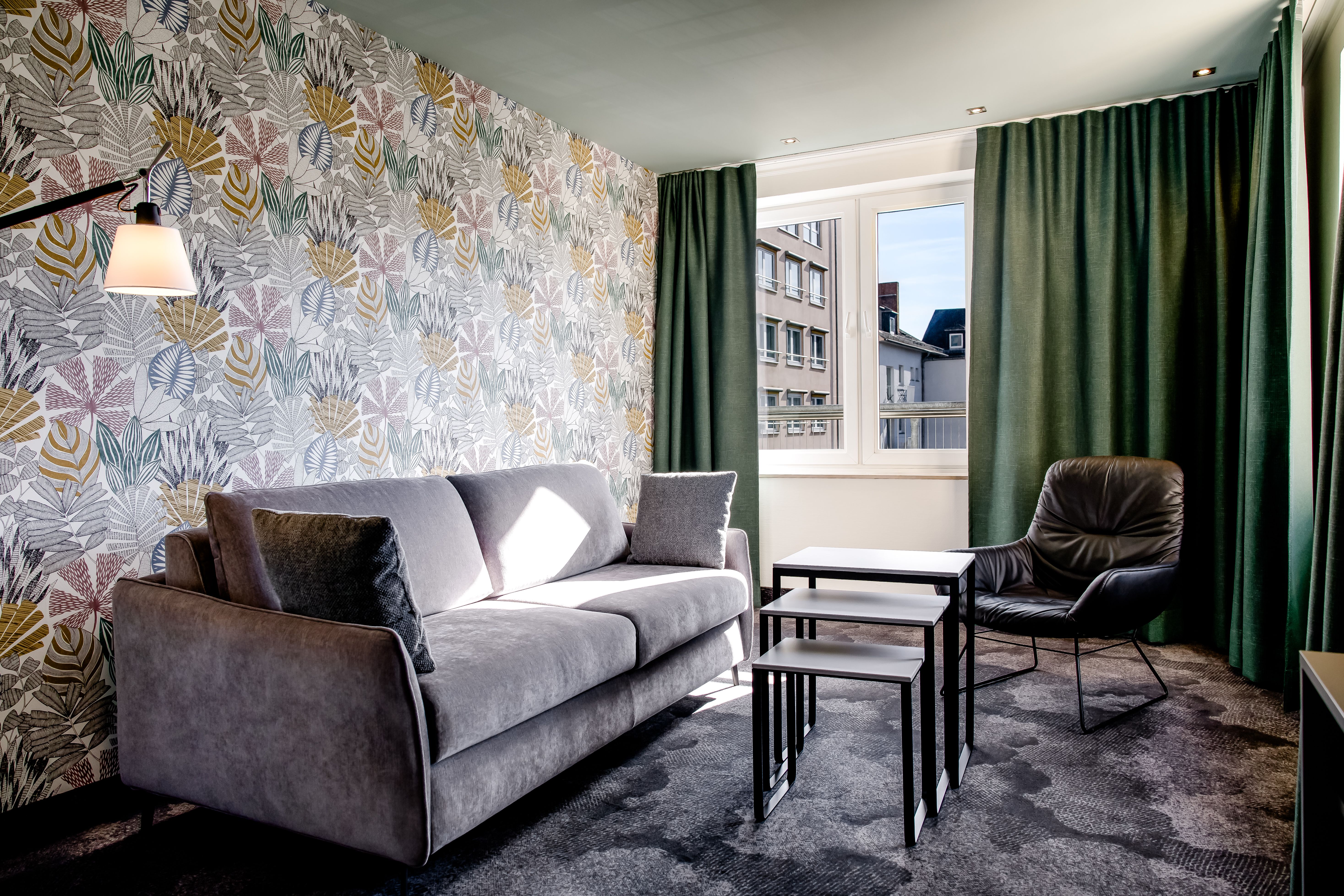 Gemütliche Sitzecke mit grauem Sofa, schwarzem Sessel und drei ineinander schiebbaren Tischen vor Fenster mit grünem Vorhang. Helle Wand mit floralem Muster sorgt für freundliche Atmosphäre.