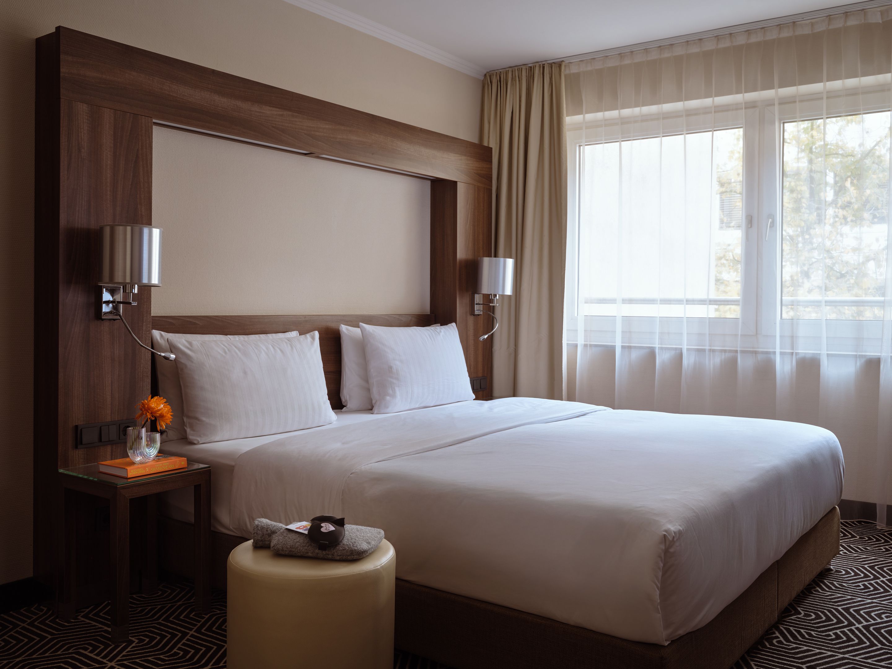 Hell eingerichtetes Hotelzimmer mit großem Doppelbett, weißen Bettwäsche, Nachttisch mit Blume und Fenster mit transparenten Vorhängen für Tageslicht.