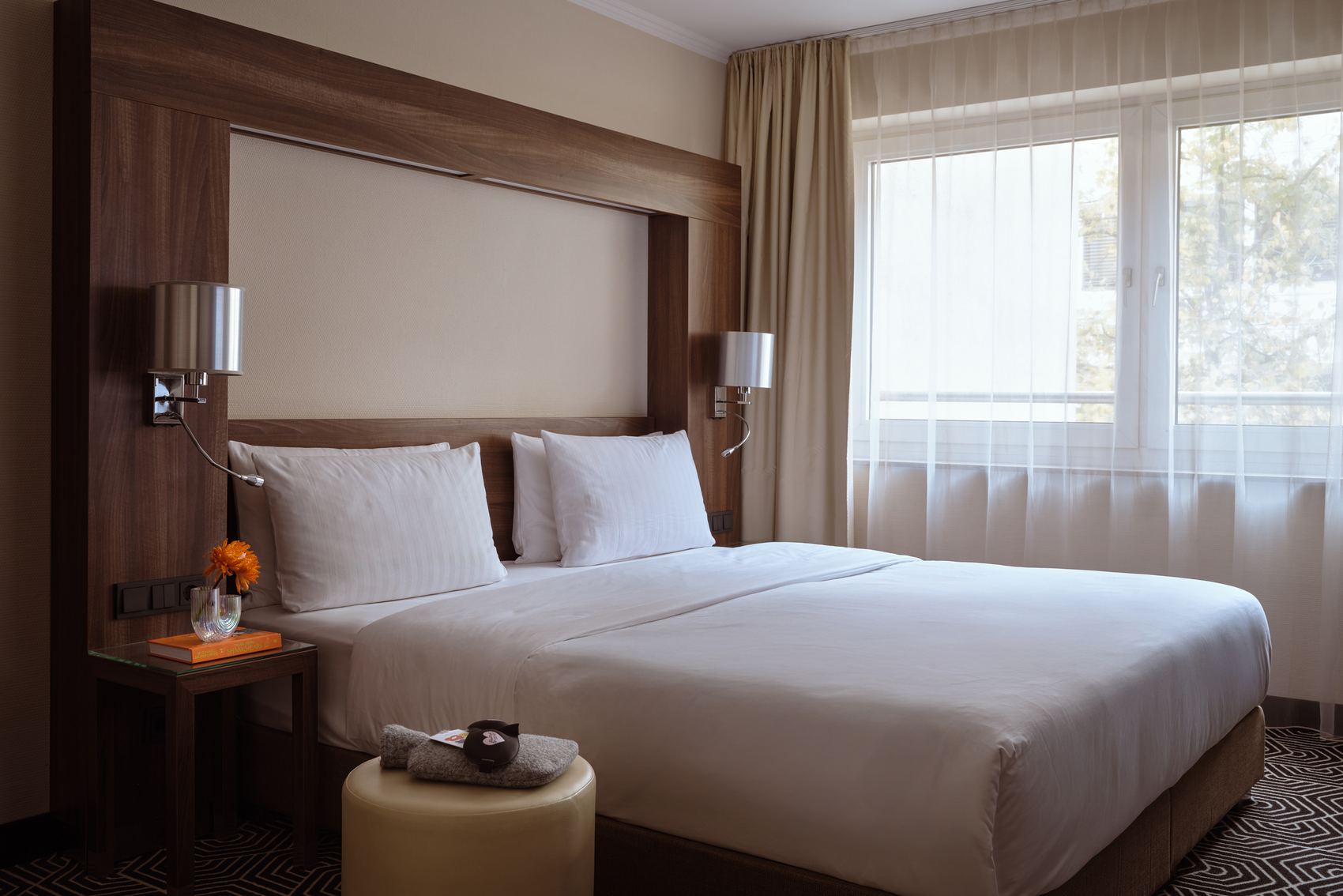 Hell eingerichtetes Hotelzimmer mit großem Doppelbett, weißen Bettwäsche, Nachttisch mit Blume und Fenster mit transparenten Vorhängen für Tageslicht.