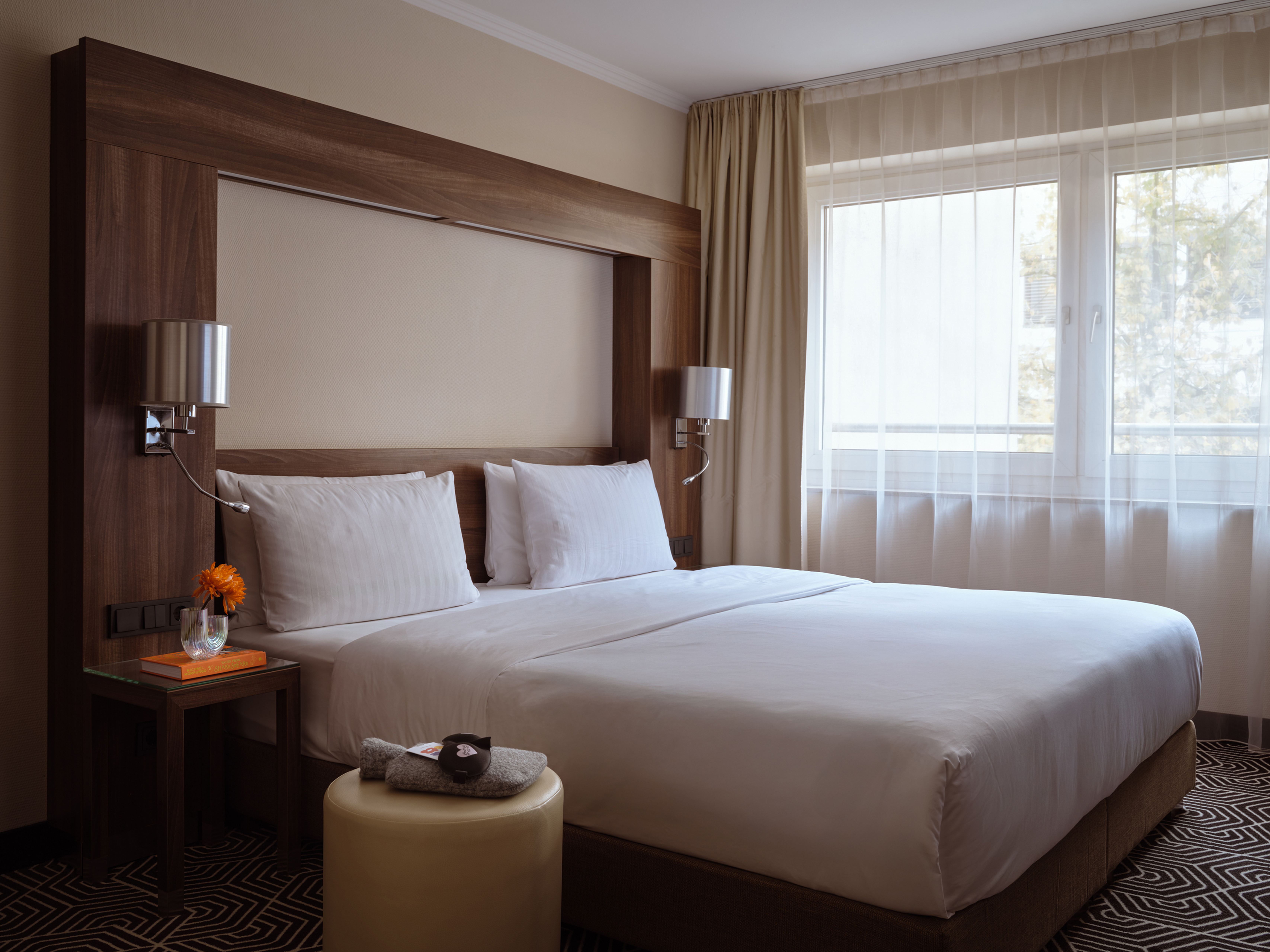 Hell eingerichtetes Hotelzimmer mit großem Doppelbett, weißen Bettwäsche, Nachttisch mit Blume und Fenster mit transparenten Vorhängen für Tageslicht. Hell eingerichtetes Hotelzimmer mit großem Doppelbett, weißen Bettwäsche, Nachttisch mit Blume und Fenster mit transparenten Vorhängen für Tageslicht.