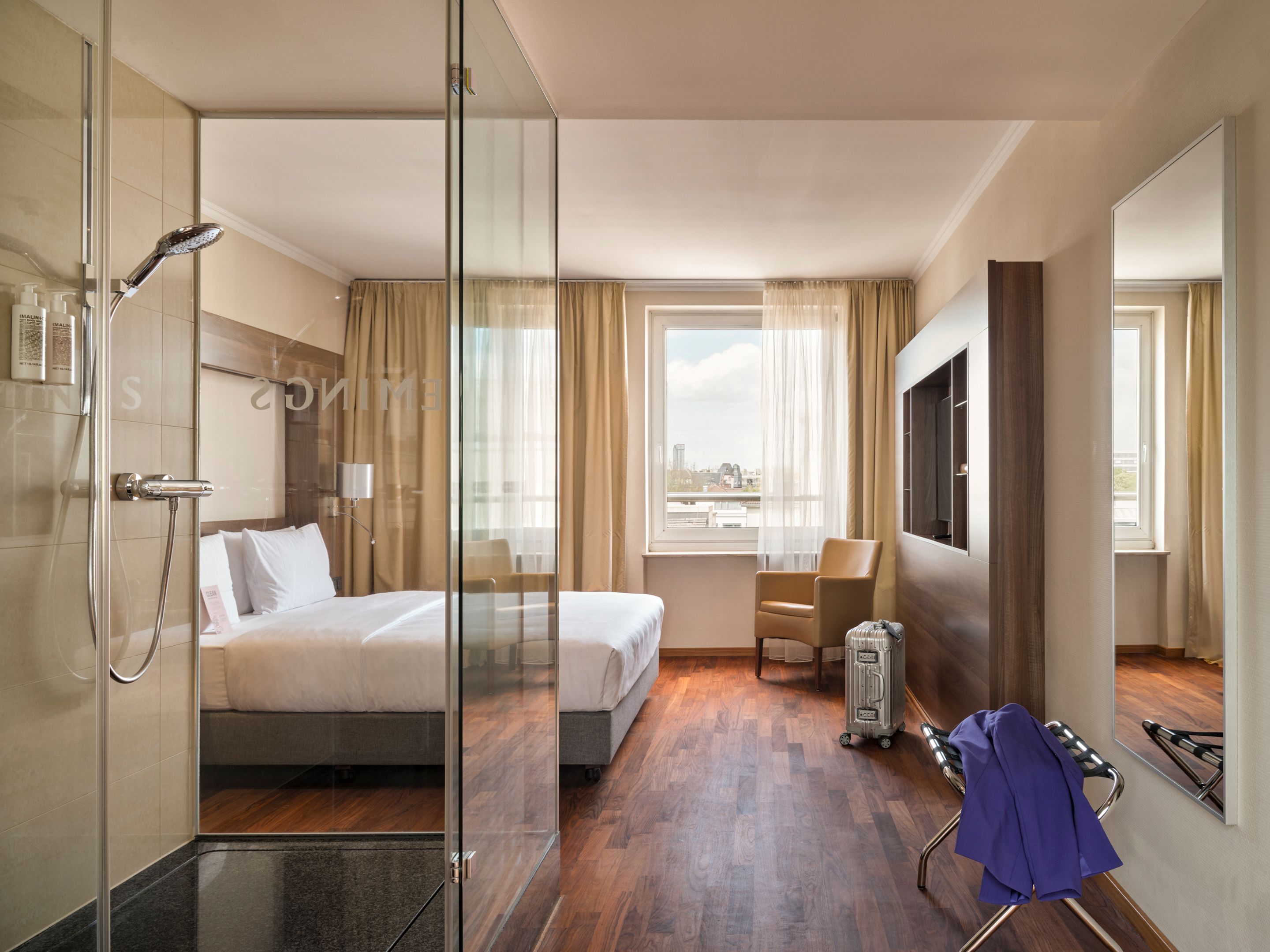 Modernes Hotelzimmer mit großem Bett, Glasdusche, Holzfußboden und Tageslicht. Helle Vorhänge, bequemer Sessel am Fenster mit Stadtblick, minimalistische Einrichtung für entspannten Aufenthalt.